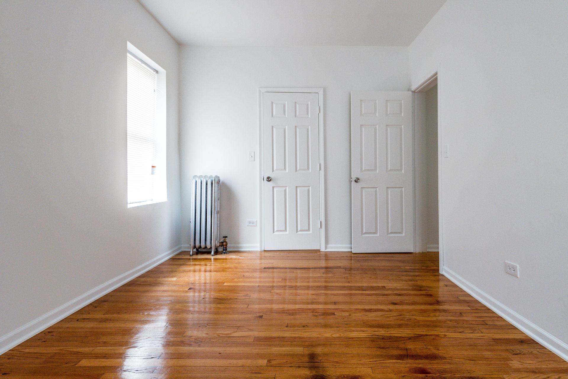 Empty room with hardwood floors, white walls, two doors, and a window with a radiator.