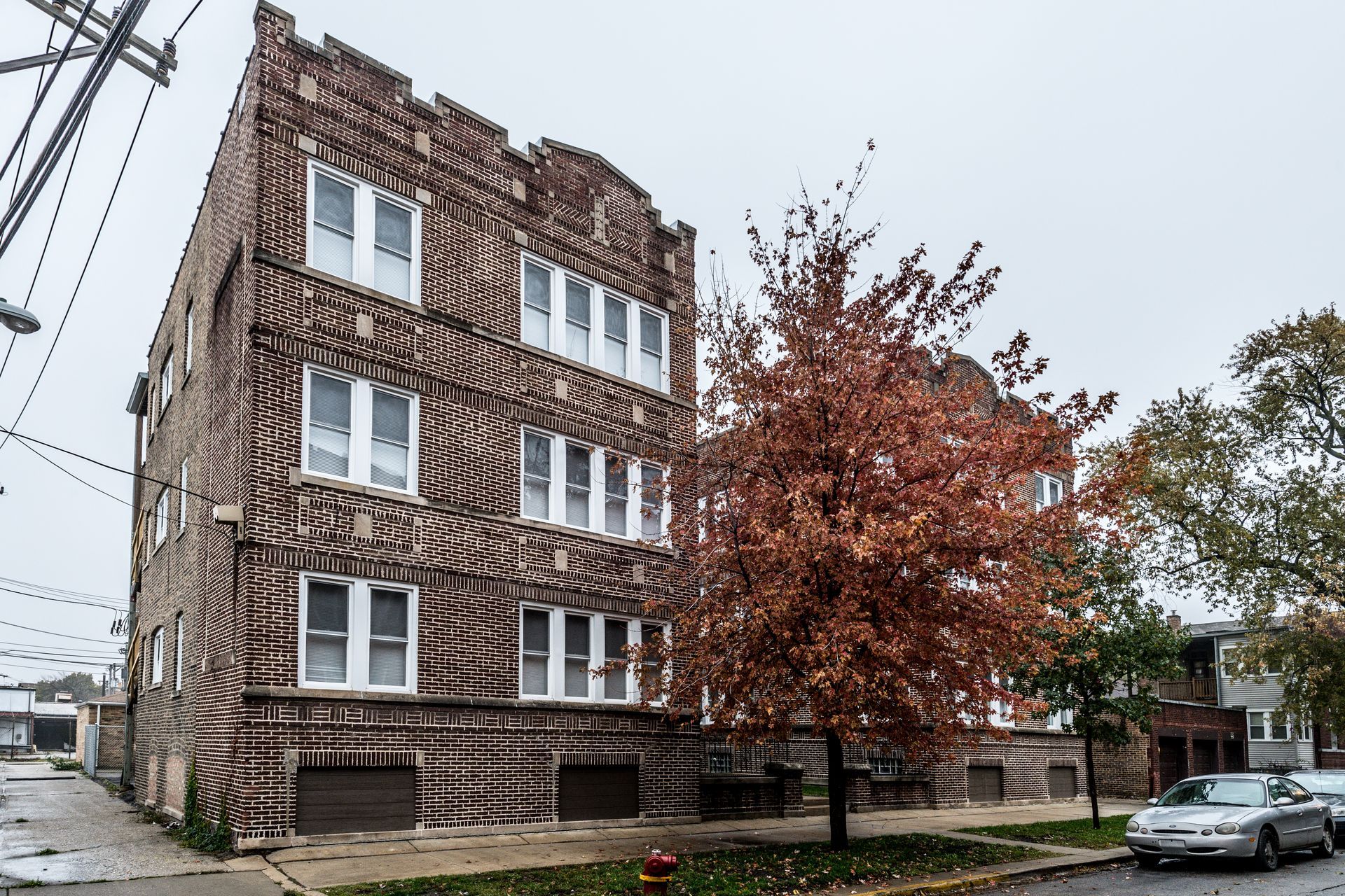 Multi-story brick building on a city street, with a red tree in front. Overcast sky.