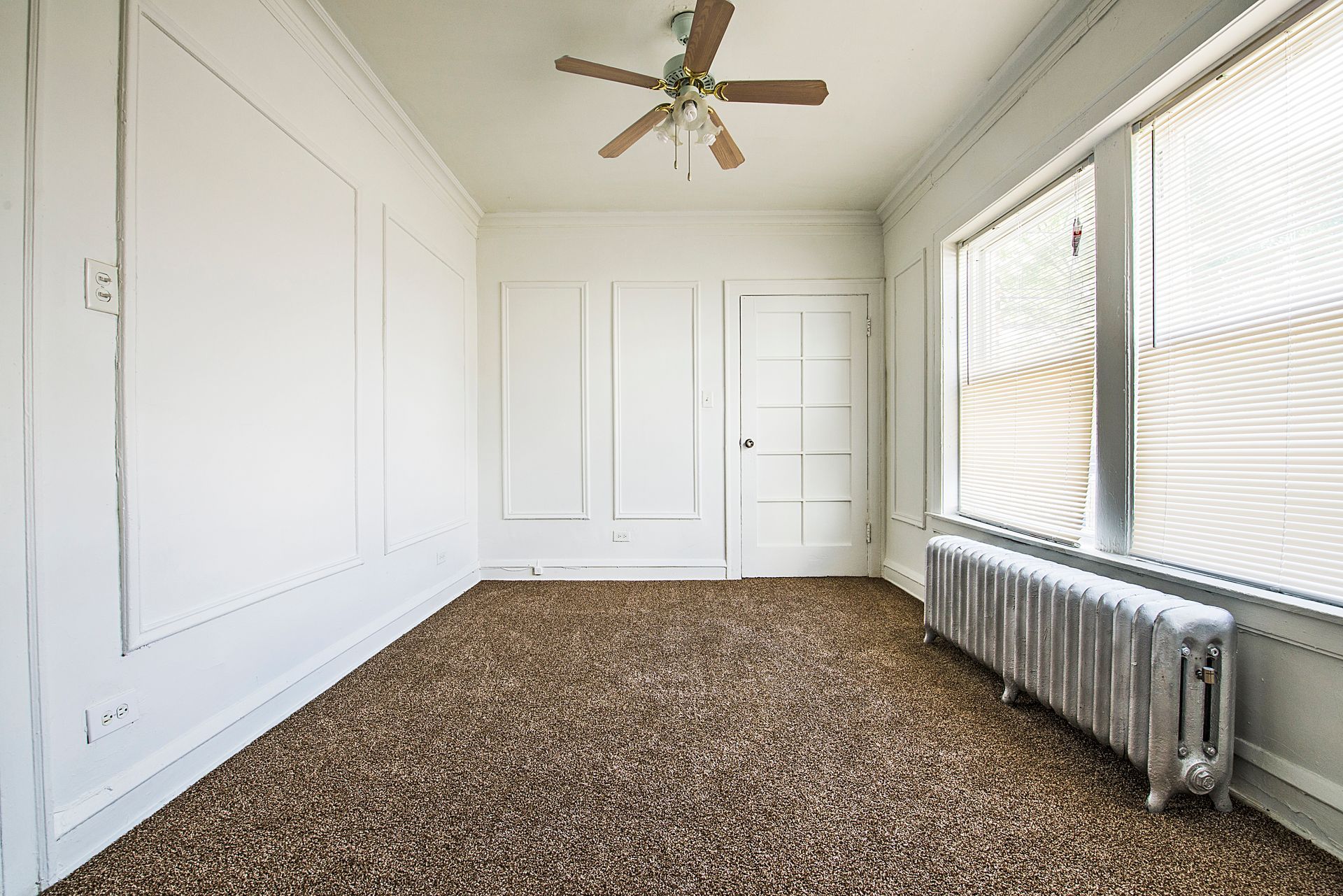 Empty room with brown carpet, white walls, large window with blinds, and ceiling fan.