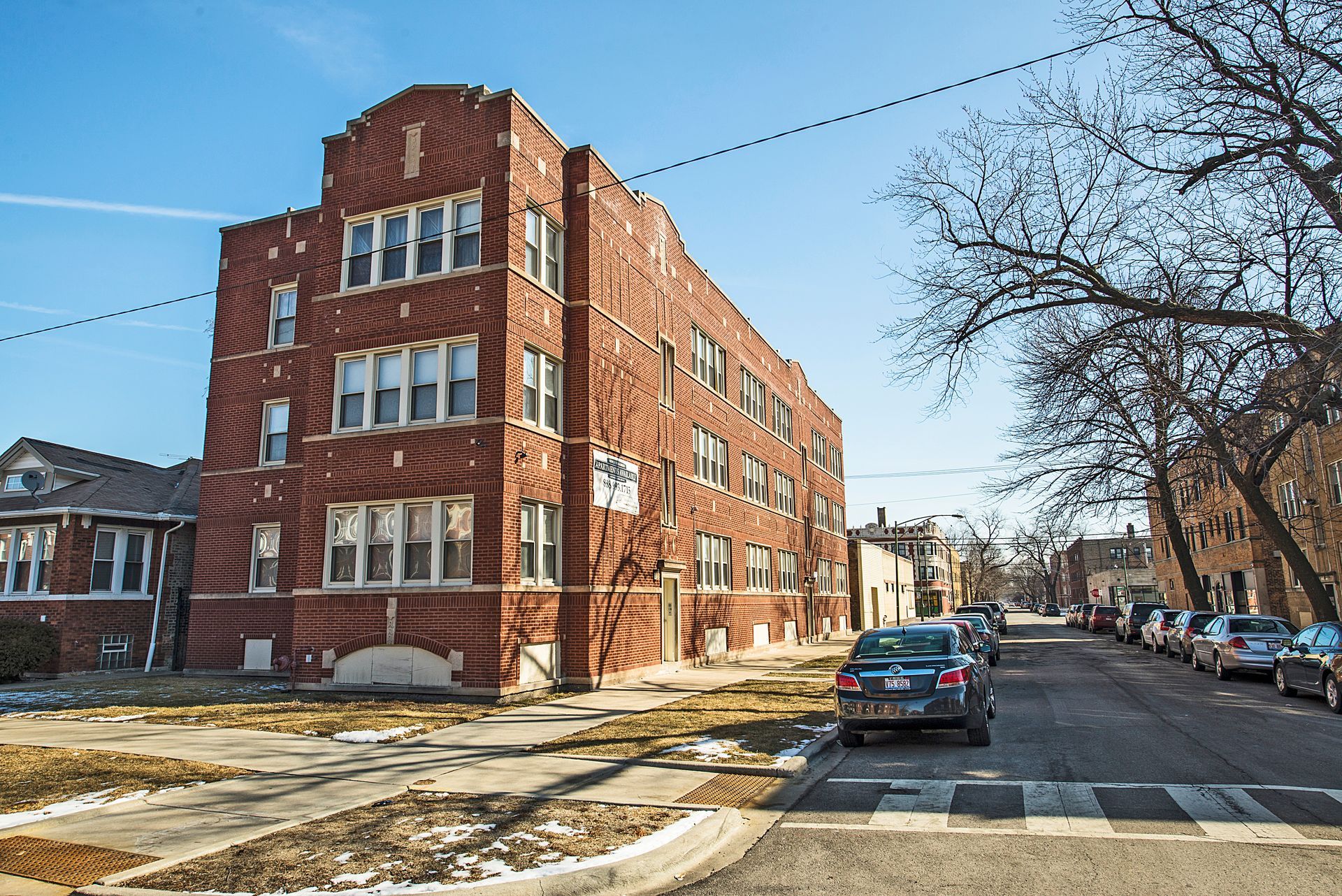 Red brick apartment building on a residential street; cars parked along the curb; sunny day.