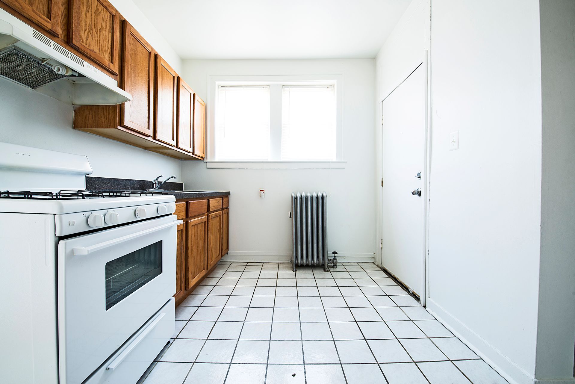 Kitchen with white tile floor, appliances, door, radiator, and window.