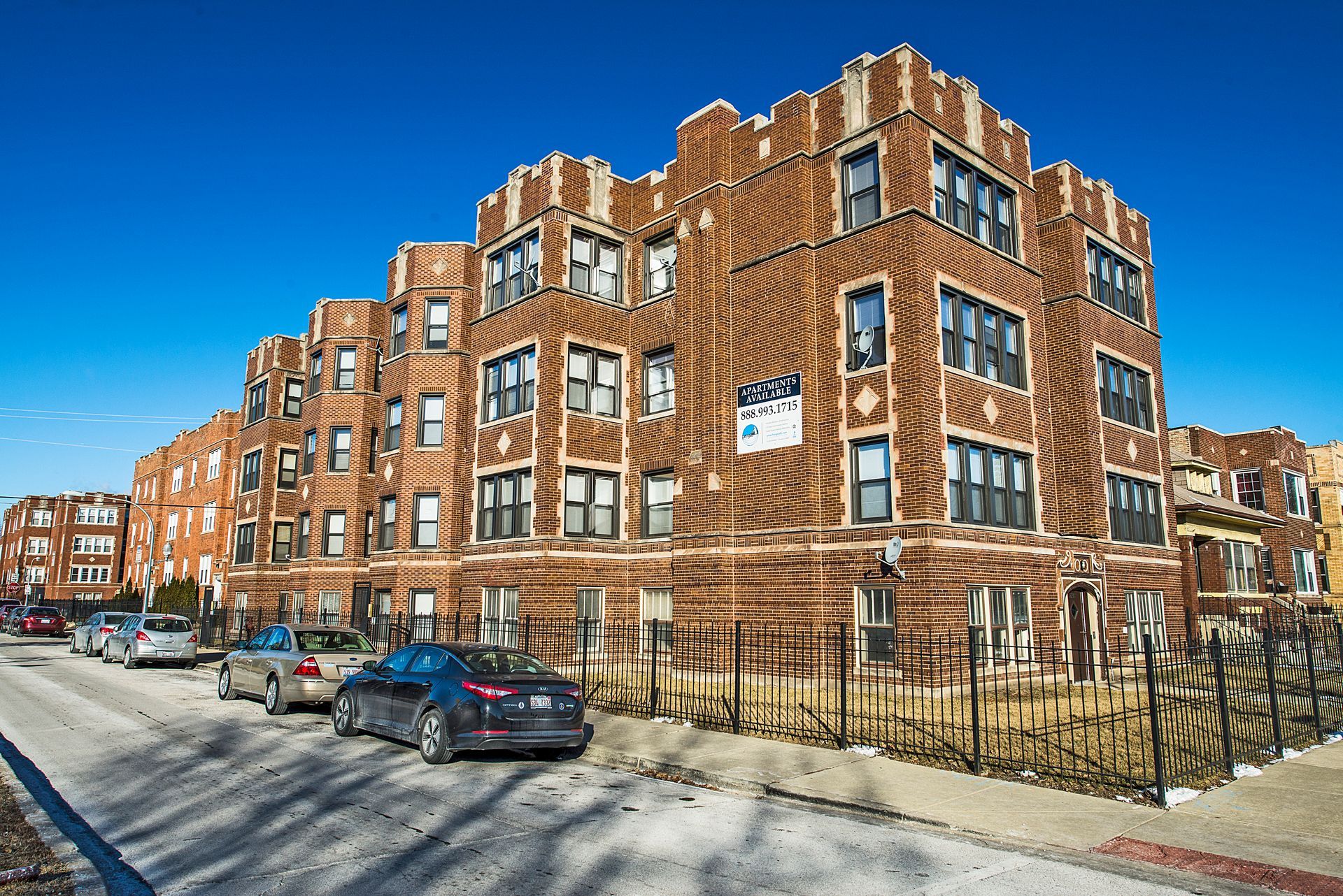 Multi-story brick apartment building on a sunny day; parked cars and a fence in front.