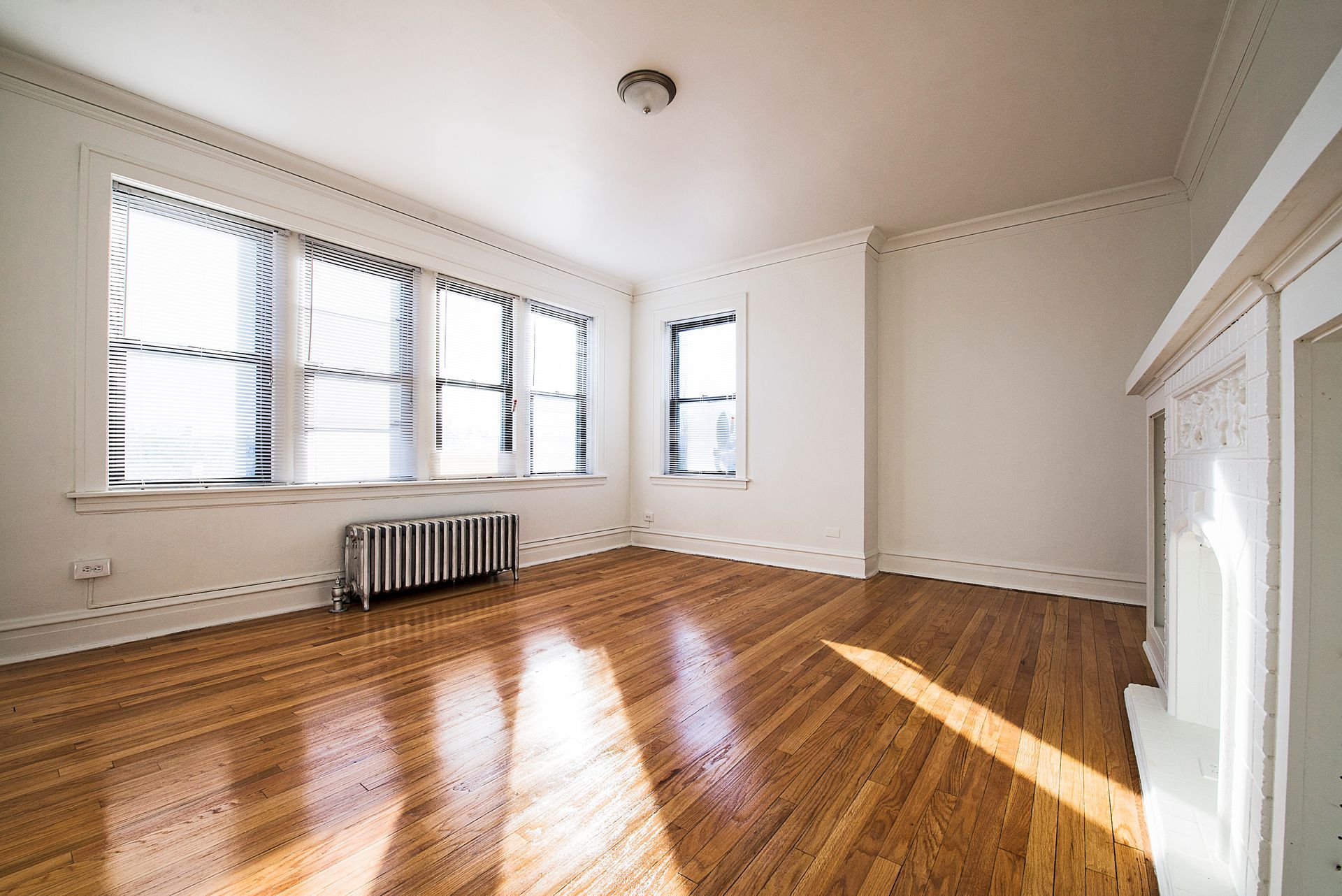 Empty room with hardwood floors, windows, and fireplace. Sunlight streams across the floor.