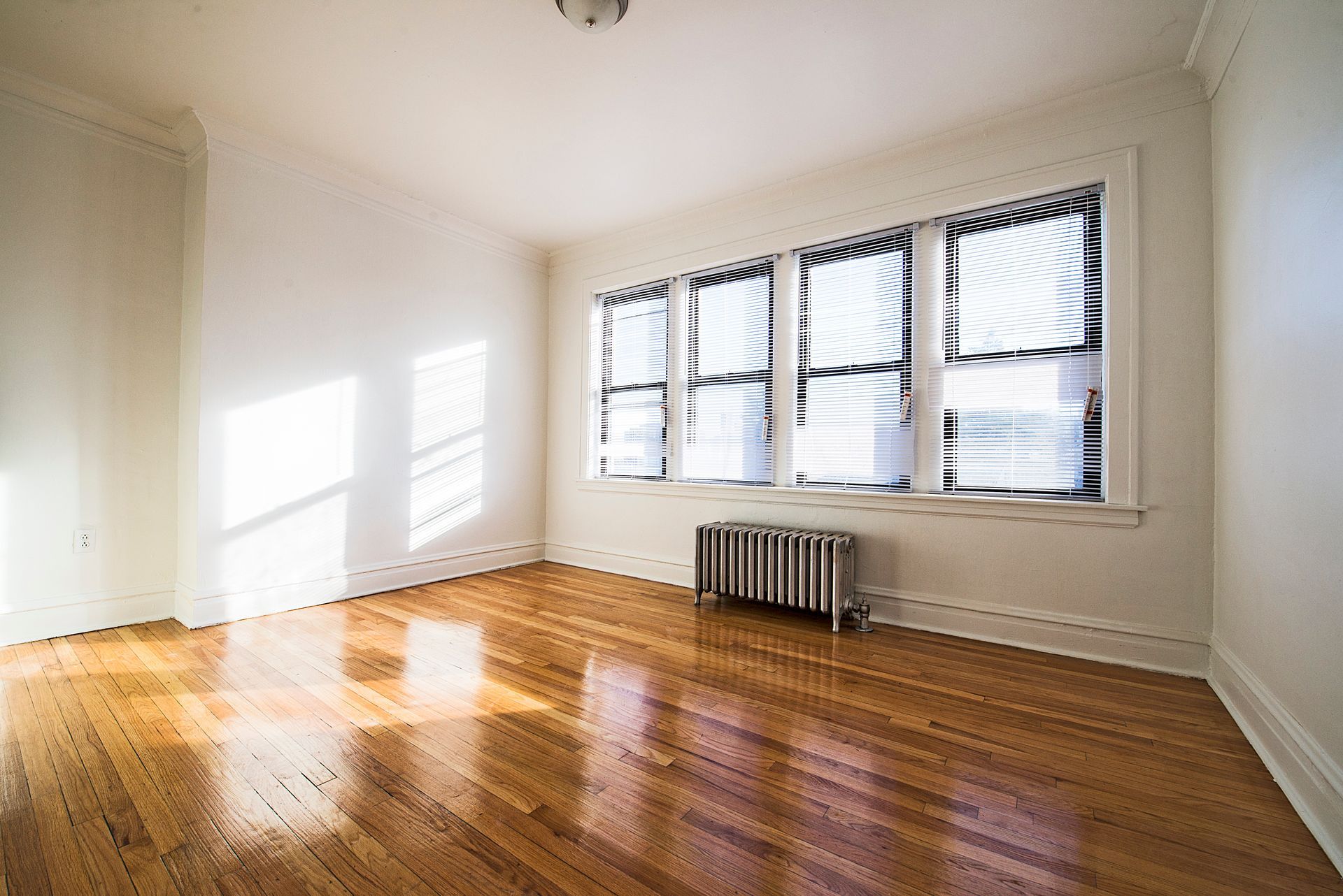 Empty room with hardwood floors, a window with blinds, and a radiator. Bright sunlight.
