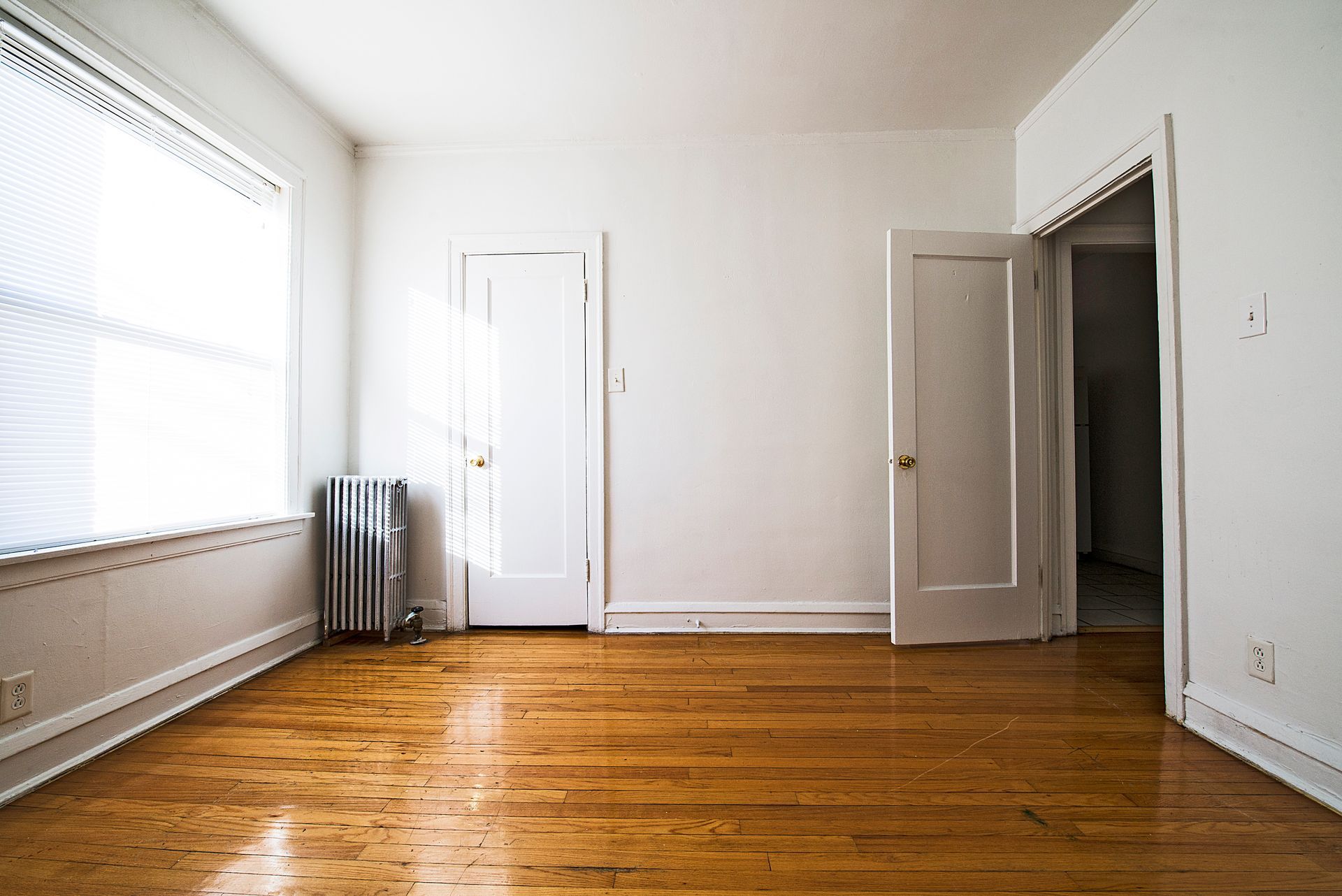 Empty room with hardwood floors, white walls, two doors, and a window with blinds.