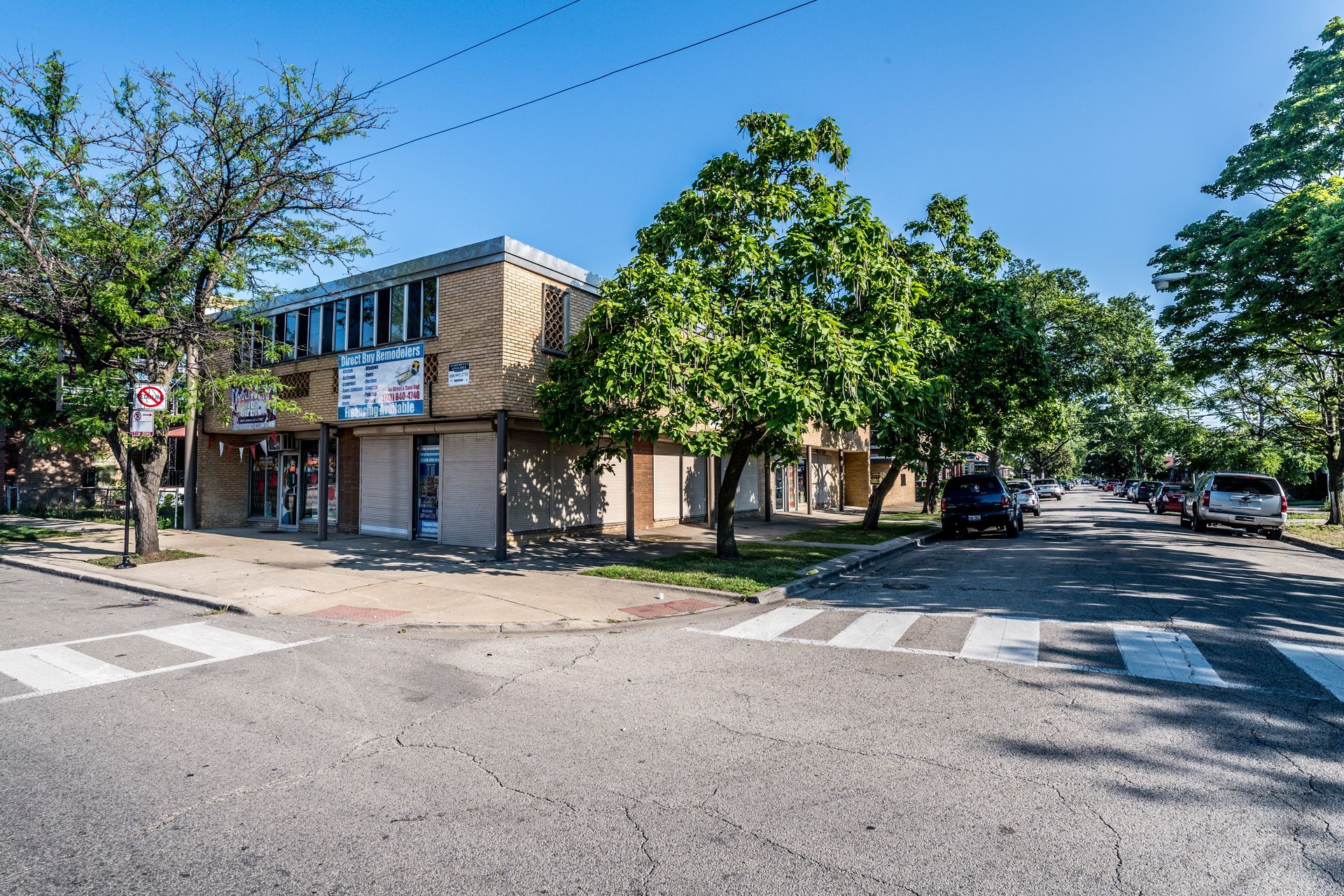 Two-story commercial building on a street corner, with crosswalks, trees, and parked cars on a sunny day.