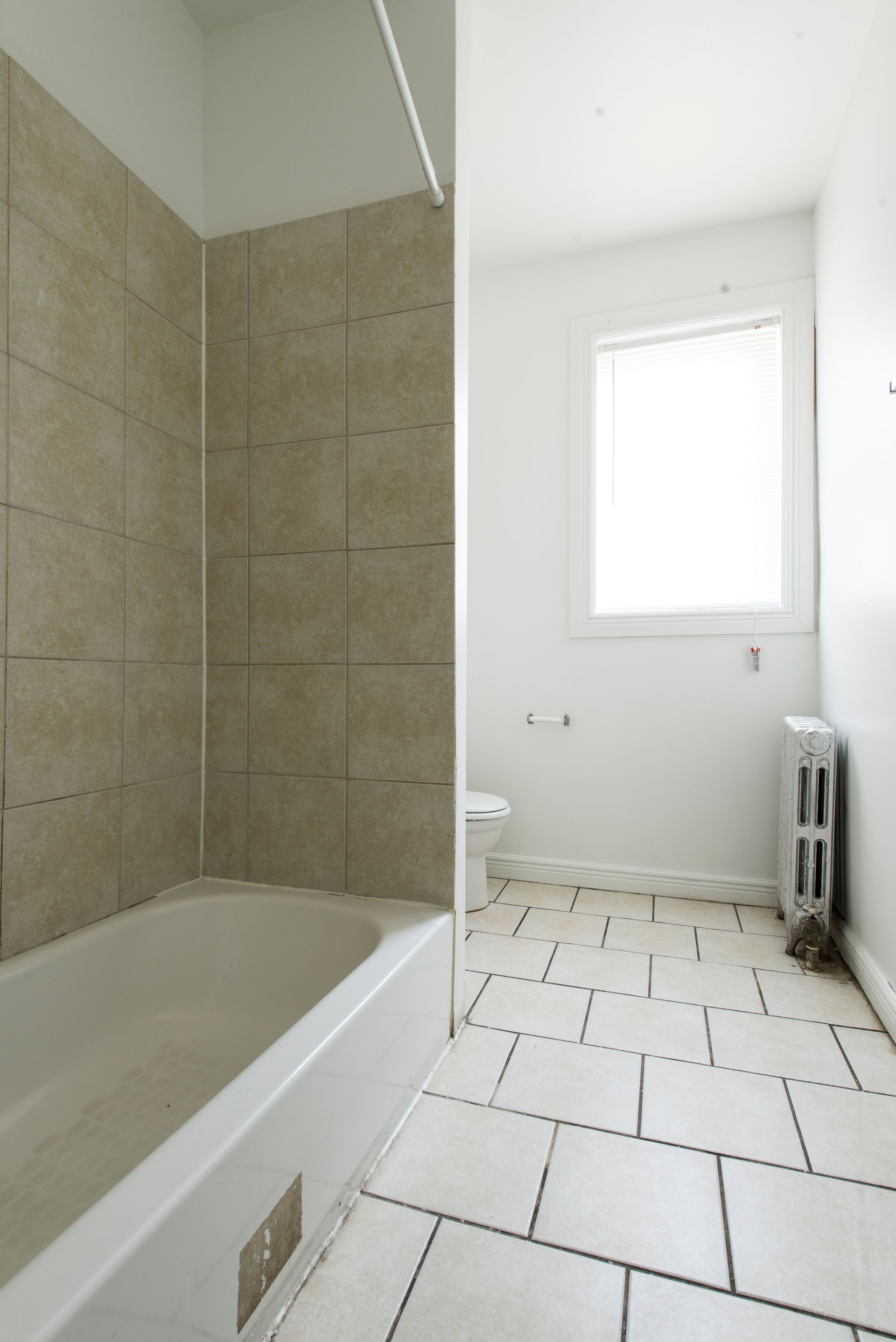 Bathroom interior: white tub, tan tile shower, white tile floor, window, radiator.
