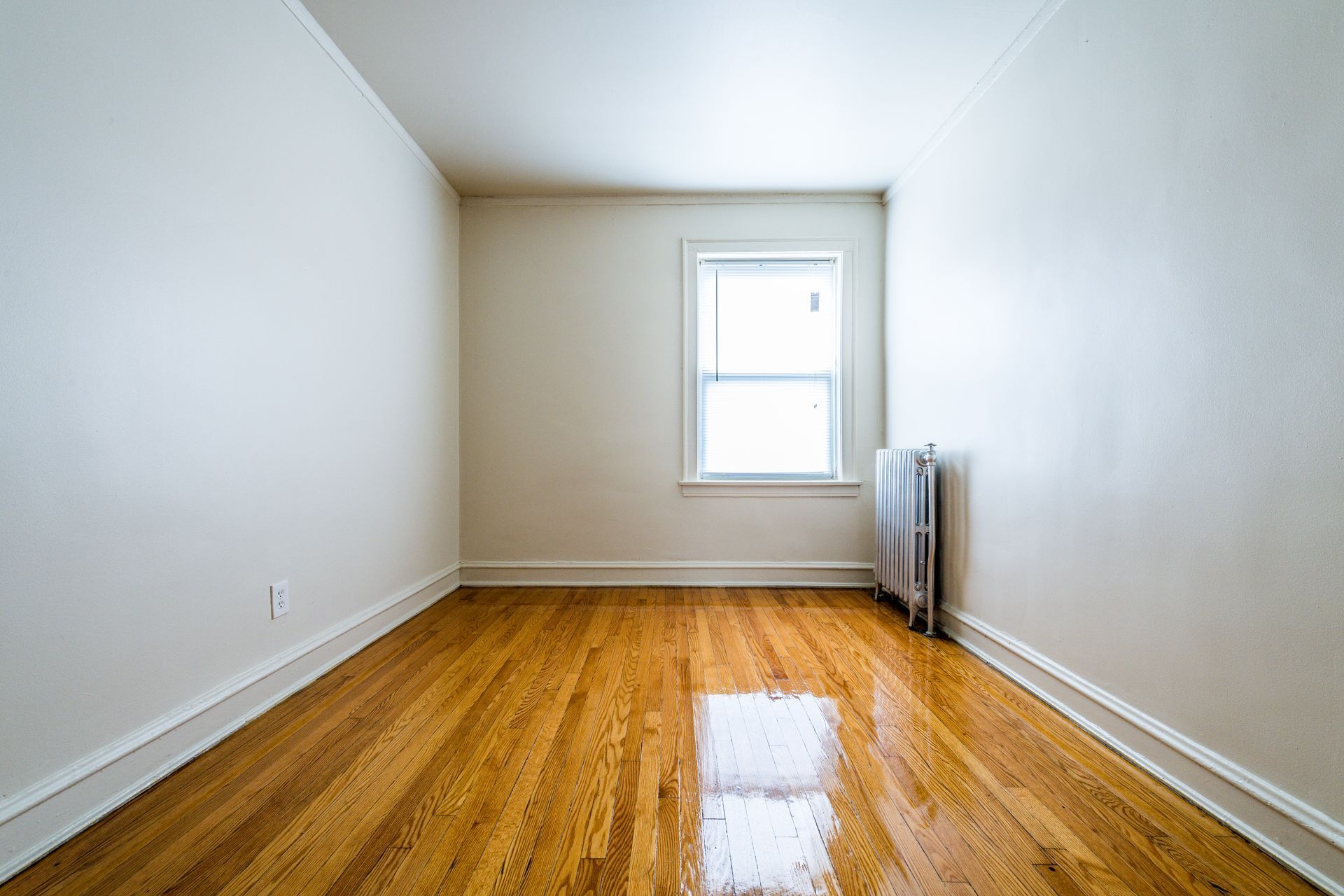 Empty room with hardwood floors, a window, and a radiator. White walls and trim.