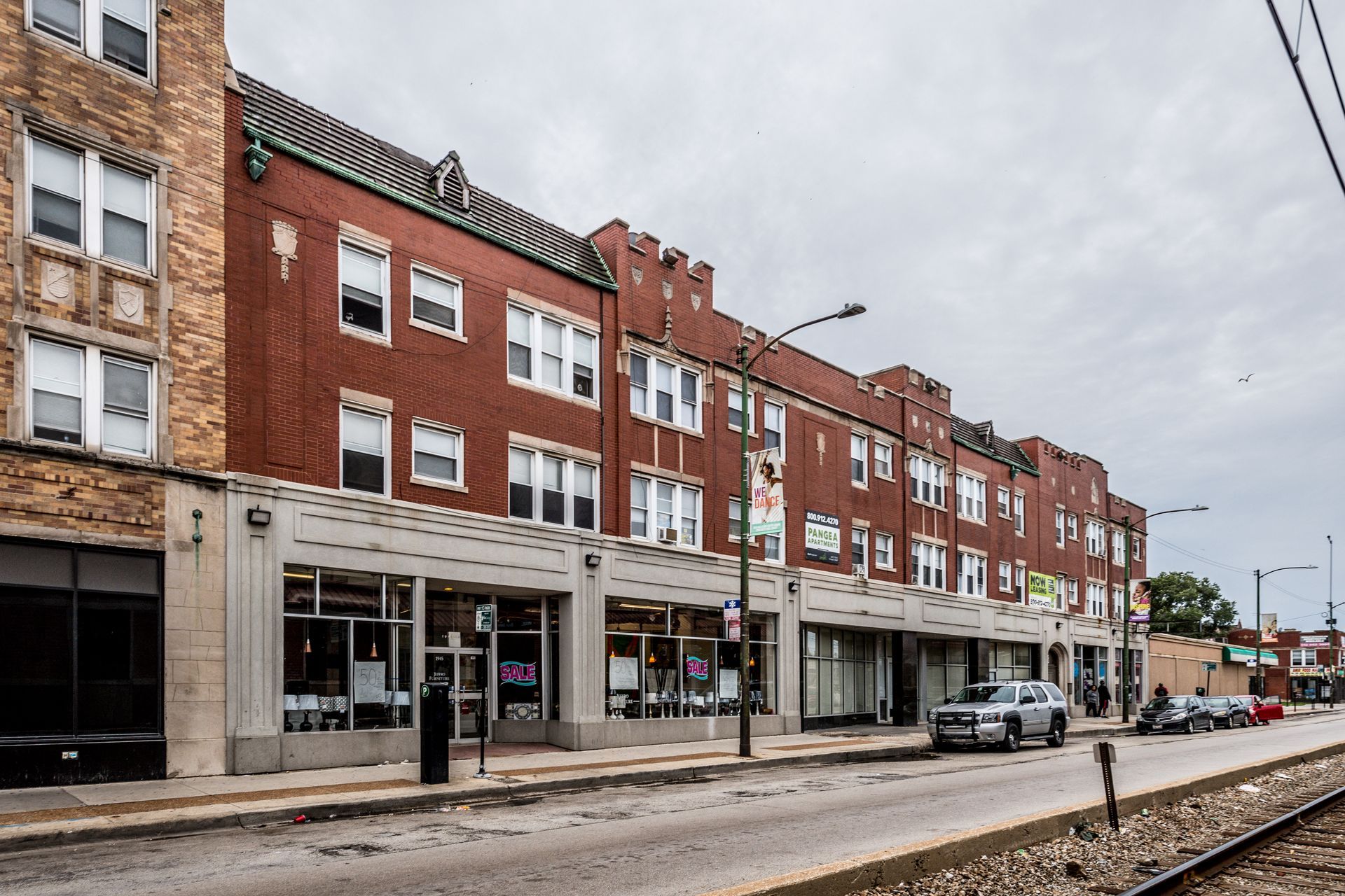 Brick buildings with storefronts on a city street, overcast sky. Cars parked near railroad tracks.