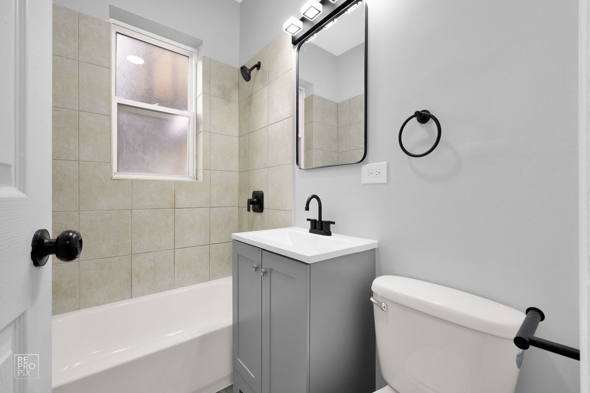Bathroom with gray vanity, white toilet, and beige tiled shower. Black fixtures against gray walls.