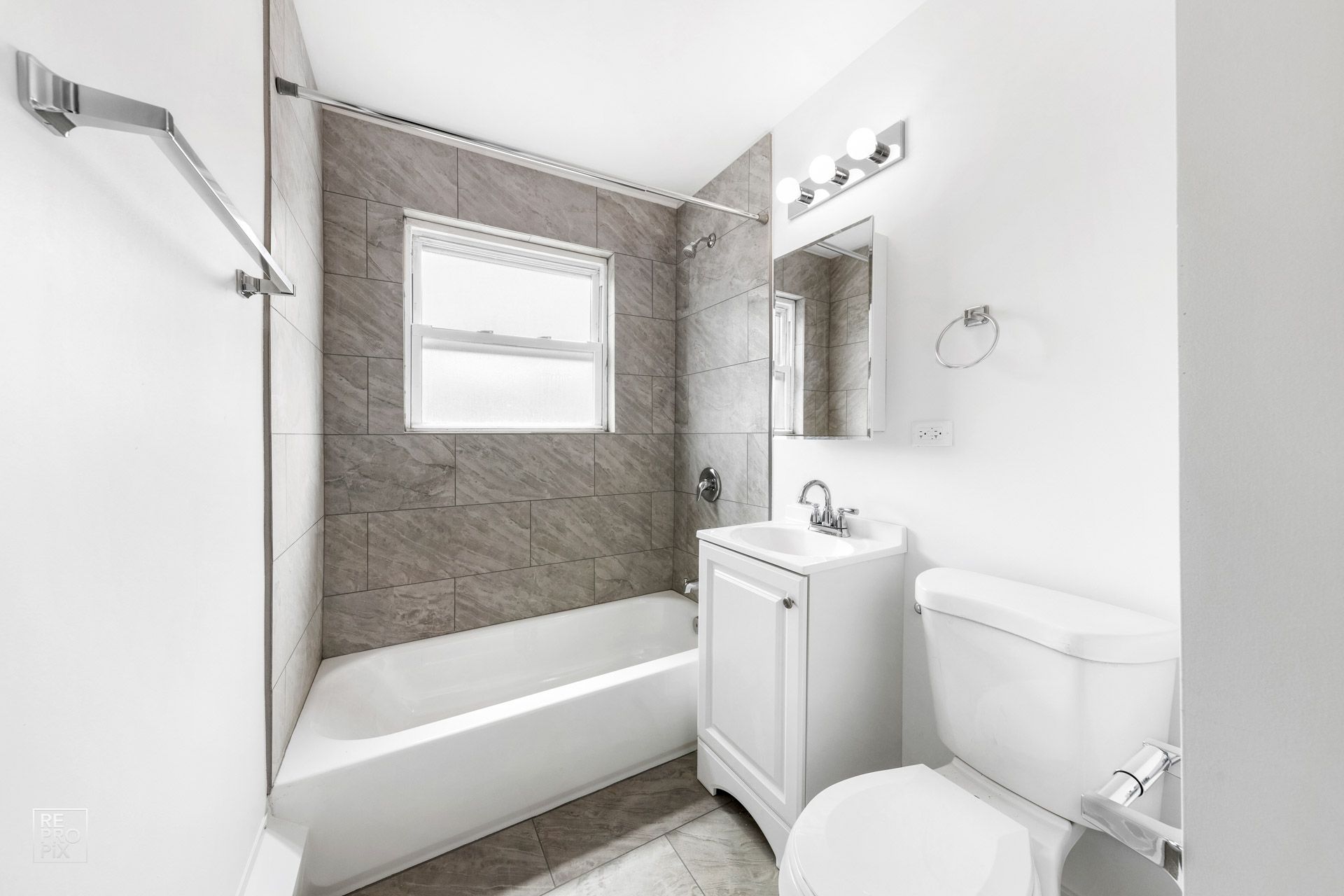 A small, bright bathroom with white fixtures, a tub/shower, and a window with gray tile walls.