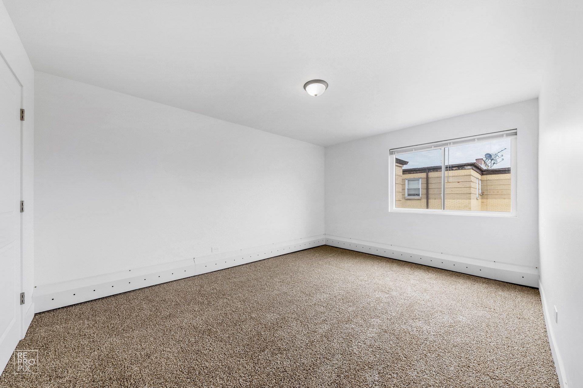 Empty room with white walls, brown carpet, window, and a single ceiling light.