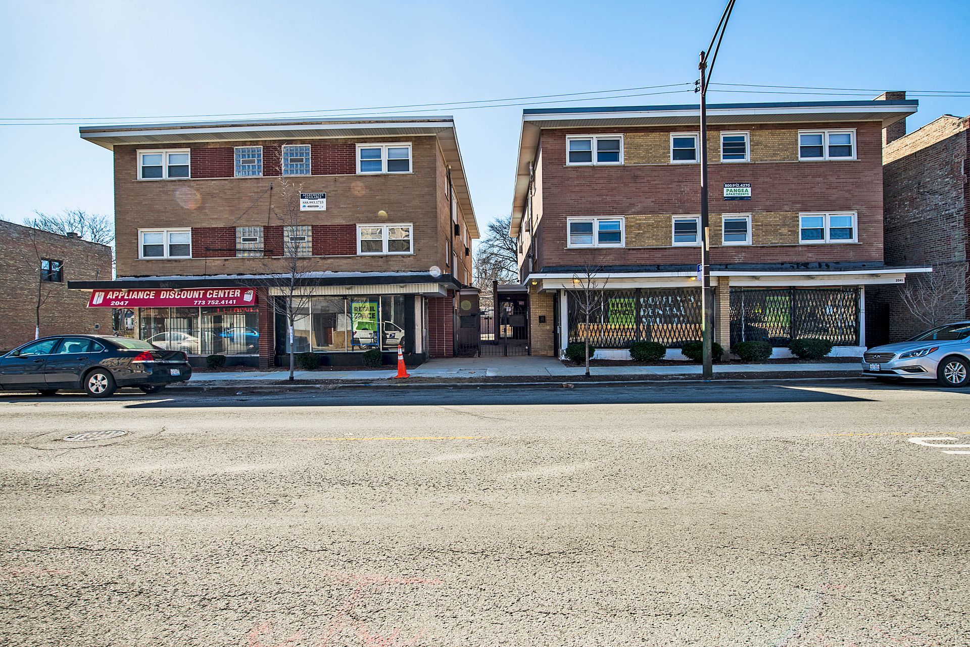 Two brick buildings with storefronts on a city street. Cars parked in front.
