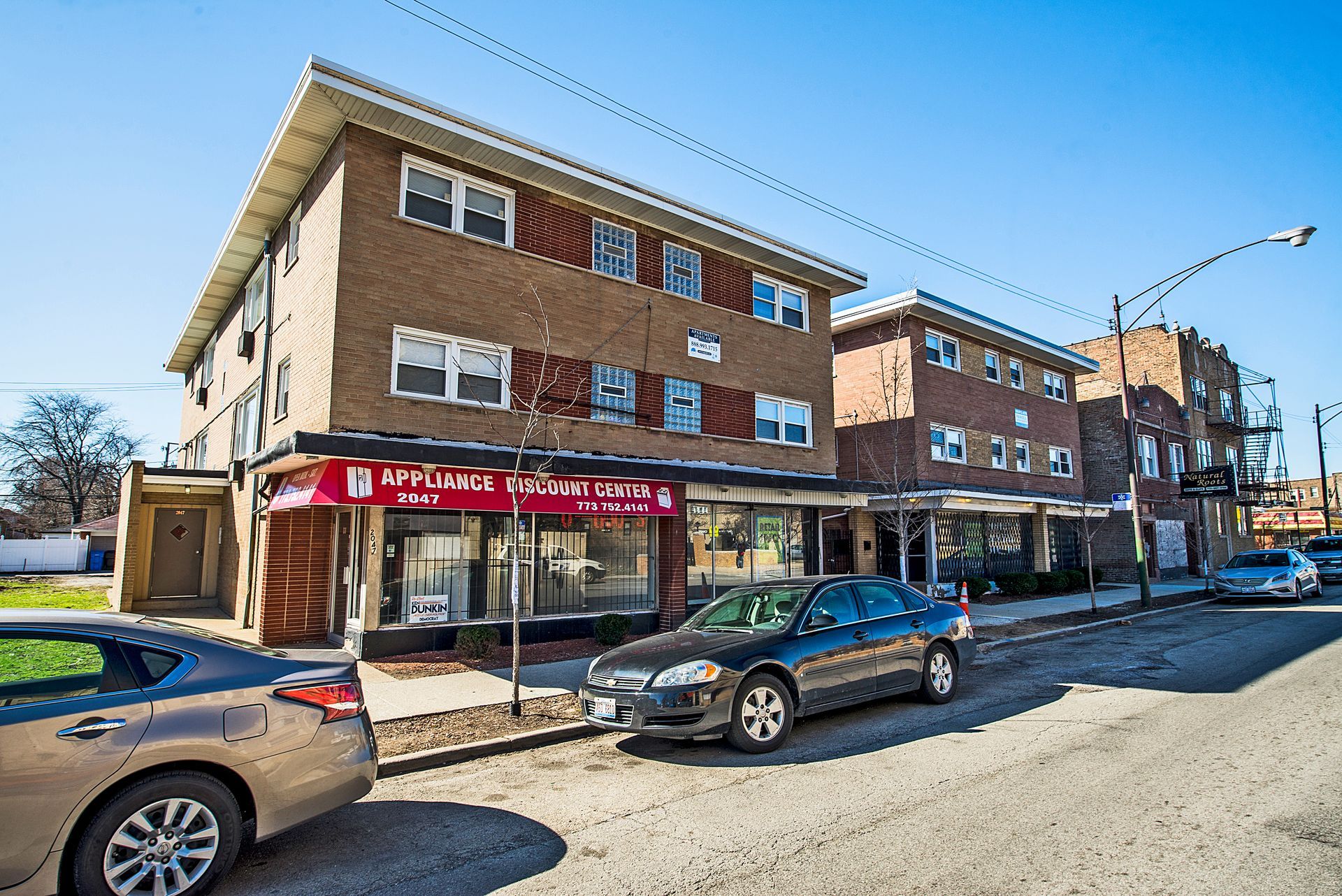 Brick buildings with storefronts along a city street; cars parked in front.