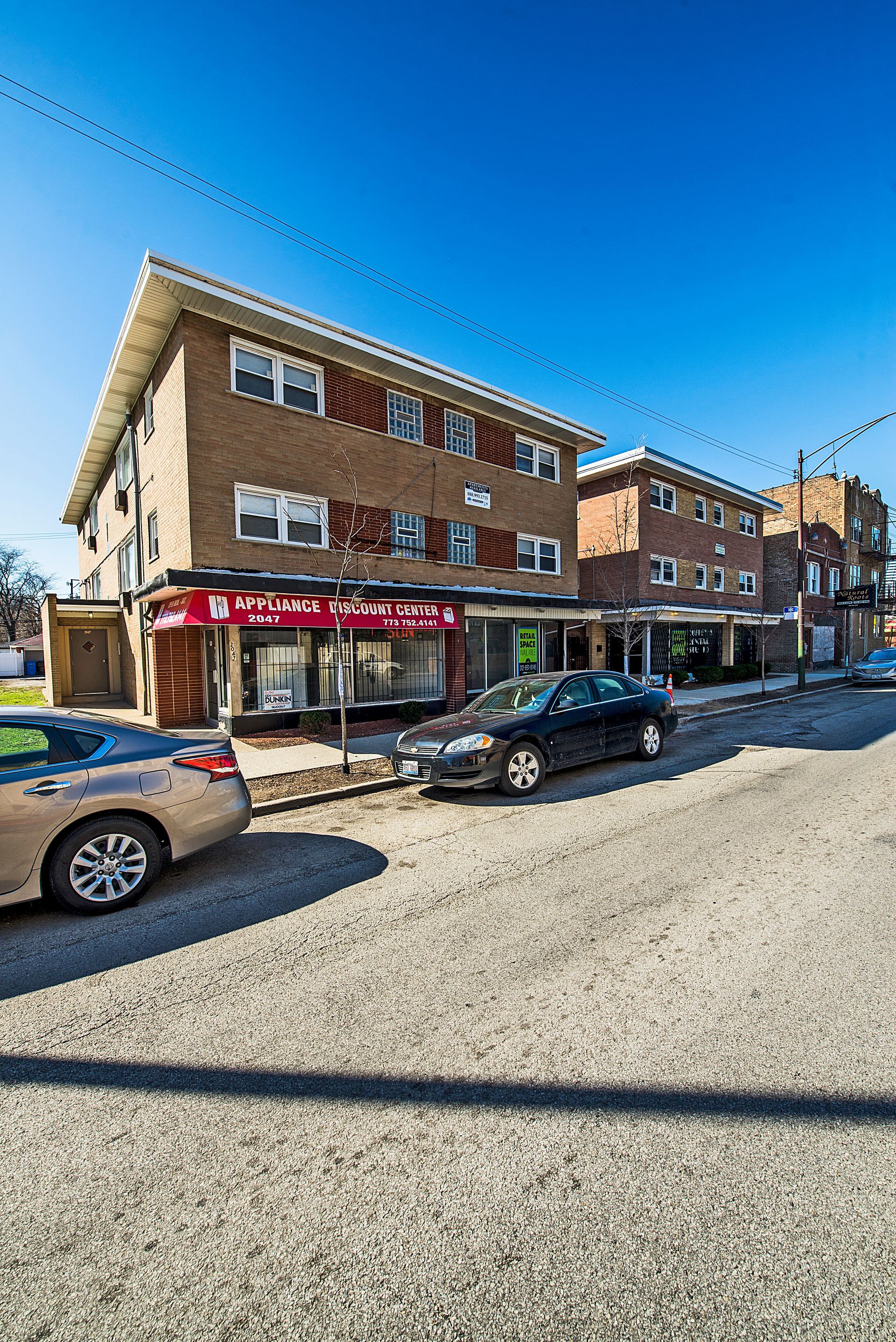 Brick building with stores at street level, cars parked on the side. Bright blue sky.