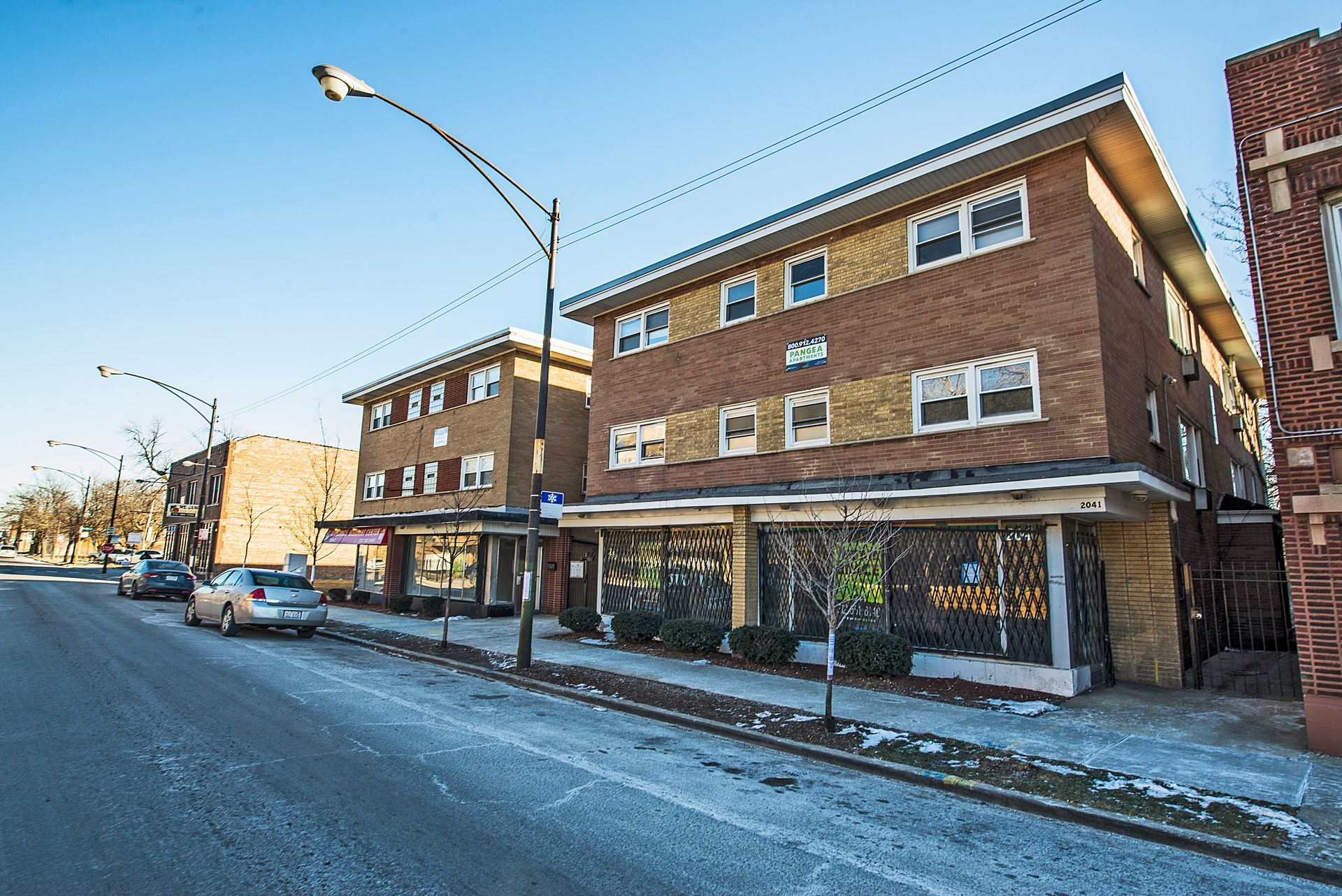 Brick apartment buildings with storefronts along a city street; cars parked on the side.