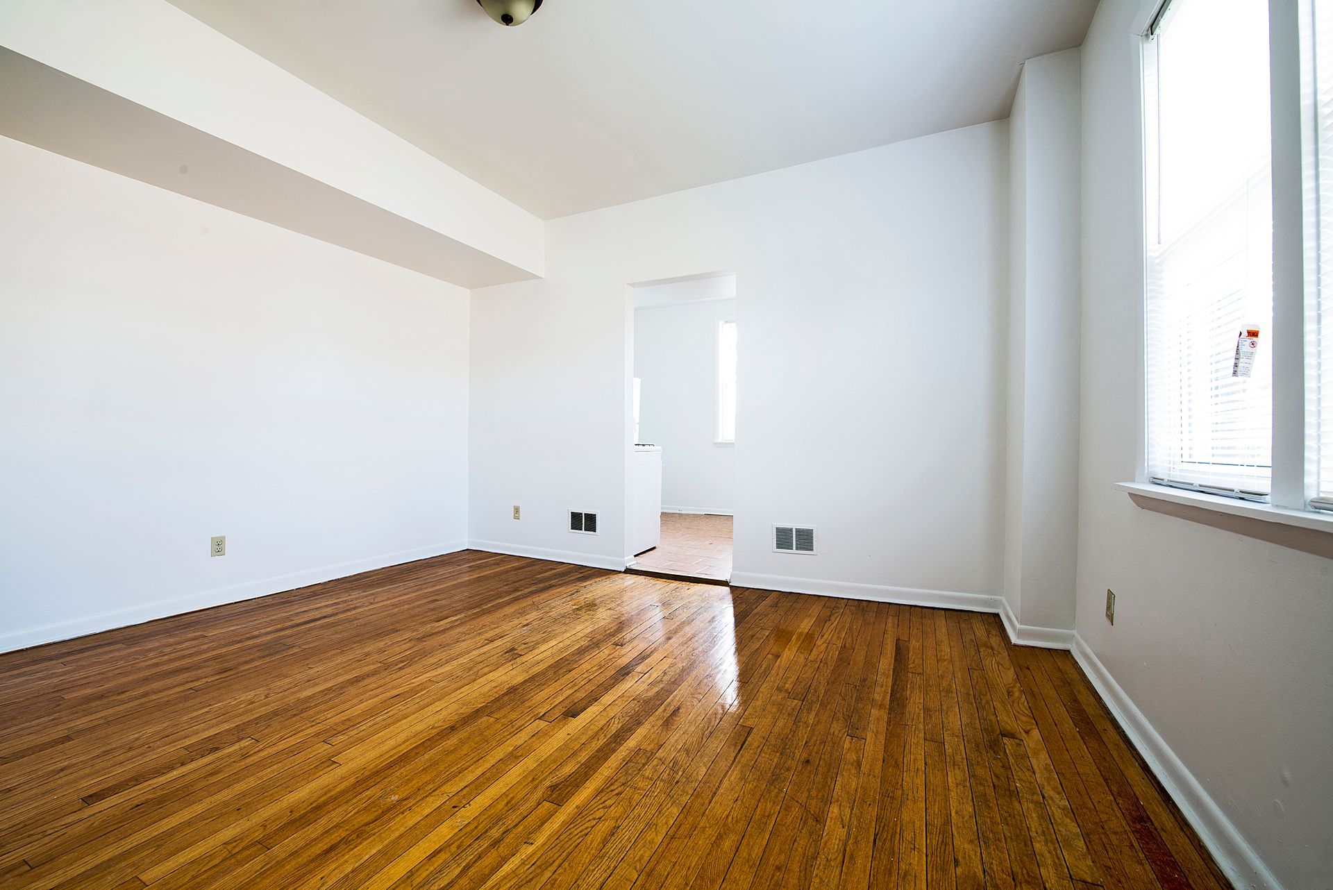 Empty room with wood floors and white walls. Window with blinds on right. Open doorway in background.
