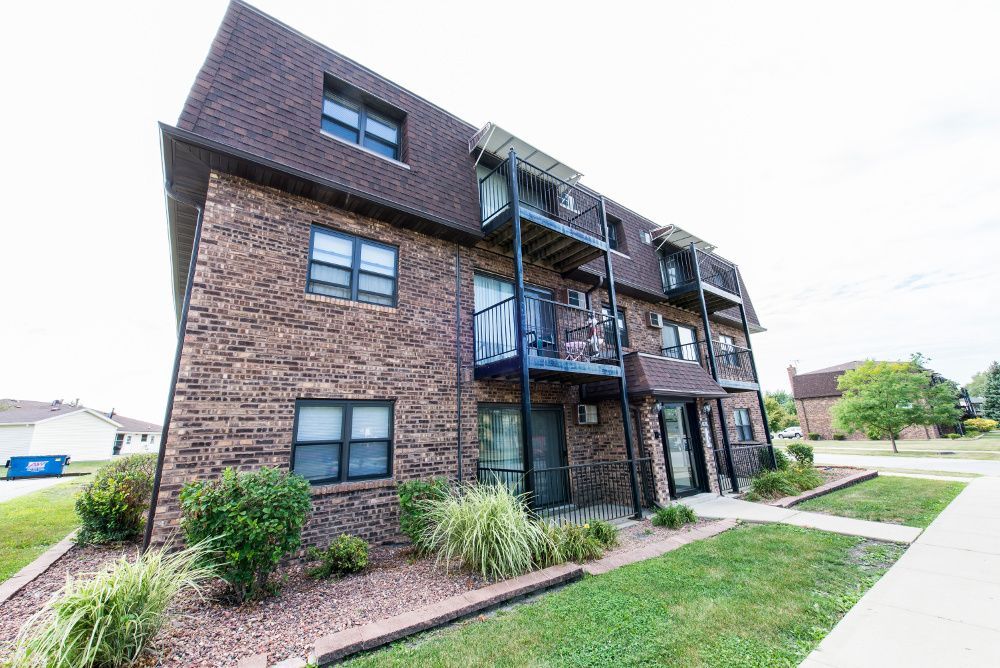 Brick apartment building with brown roof and balconies, surrounded by grass and sidewalk.