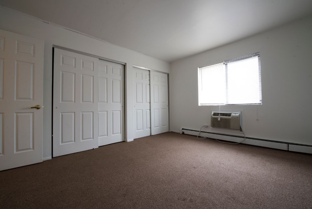 Empty bedroom with brown carpet, white walls, closet doors, and a window with an air conditioner.
