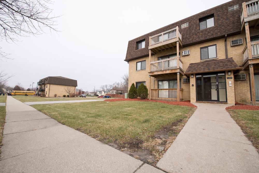 Apartment building with brown roof, tan exterior, and a sidewalk leading to the entrance. Overcast day.