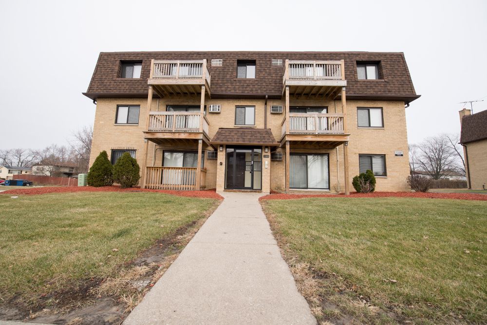 Three-story brick apartment building with brown roof and balconies, walkway leading to entrance.