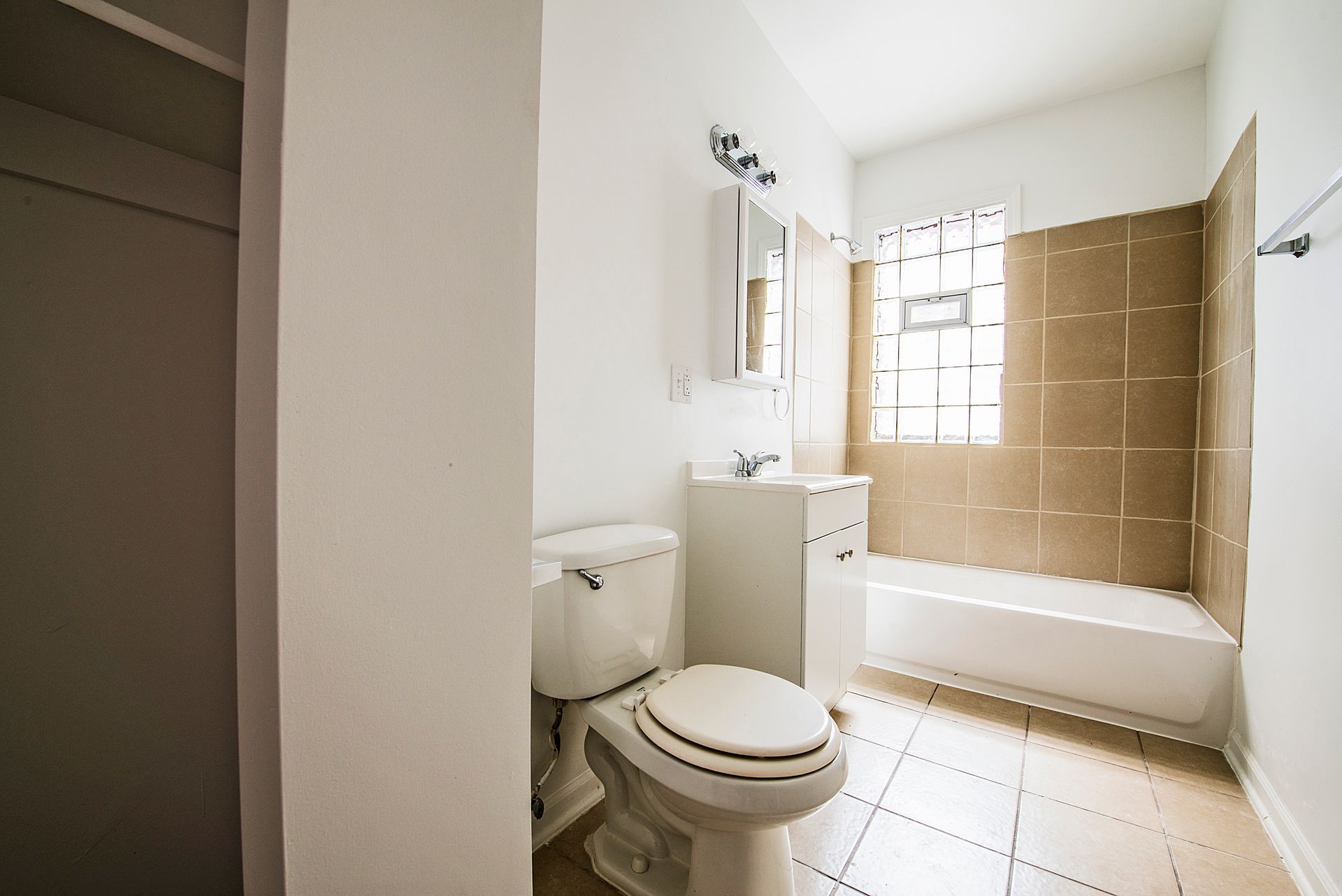 Small, white bathroom with toilet, sink, tub, and window with beige tile surround.