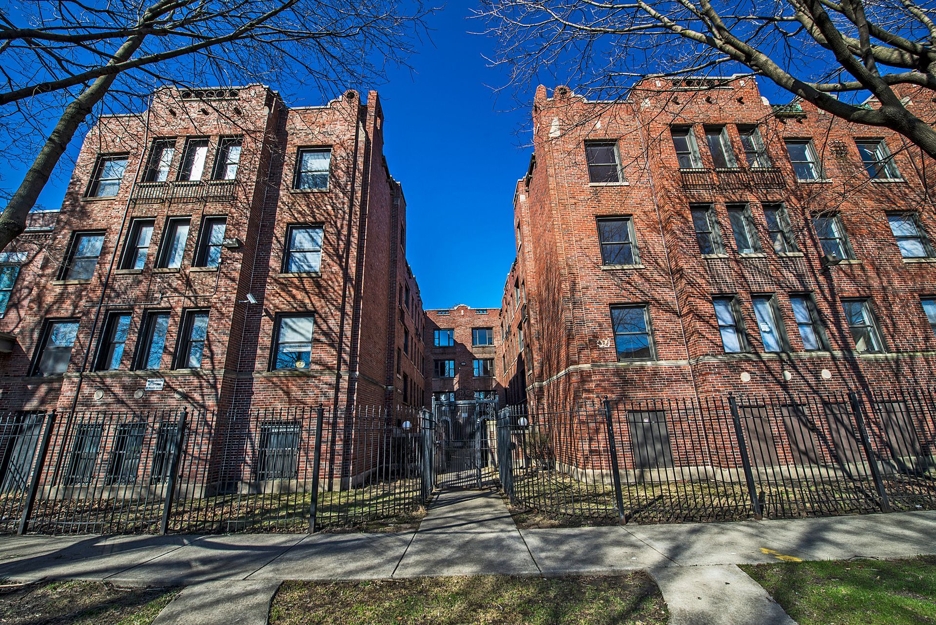 Red brick apartment buildings under a bright blue sky, with trees and a black fence in the foreground.