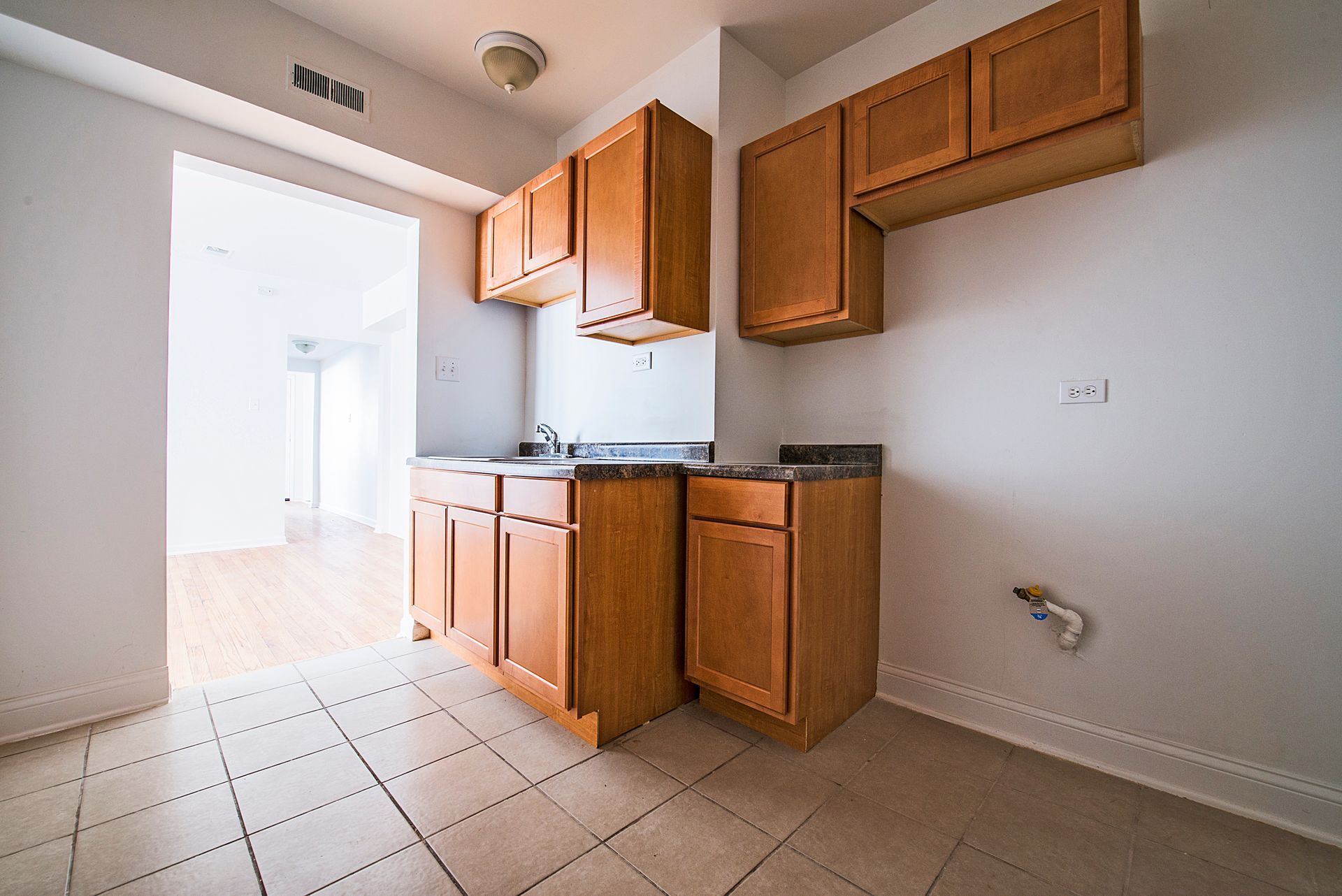 Kitchen with light brown cabinets, beige tile floor, and white walls. An open doorway leads to another room.