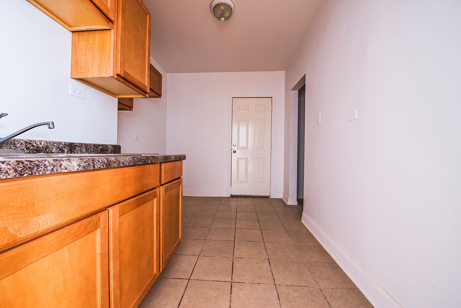 Kitchen with light-brown cabinets, gray countertop, and white walls. Tile floor and a doorway.