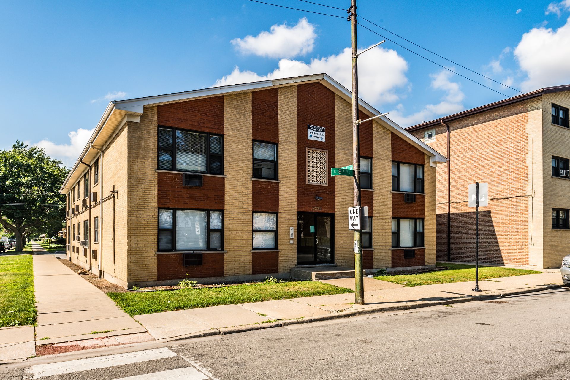 Two-story brick apartment building with brown stripes, sidewalk, and street. Sunny day.