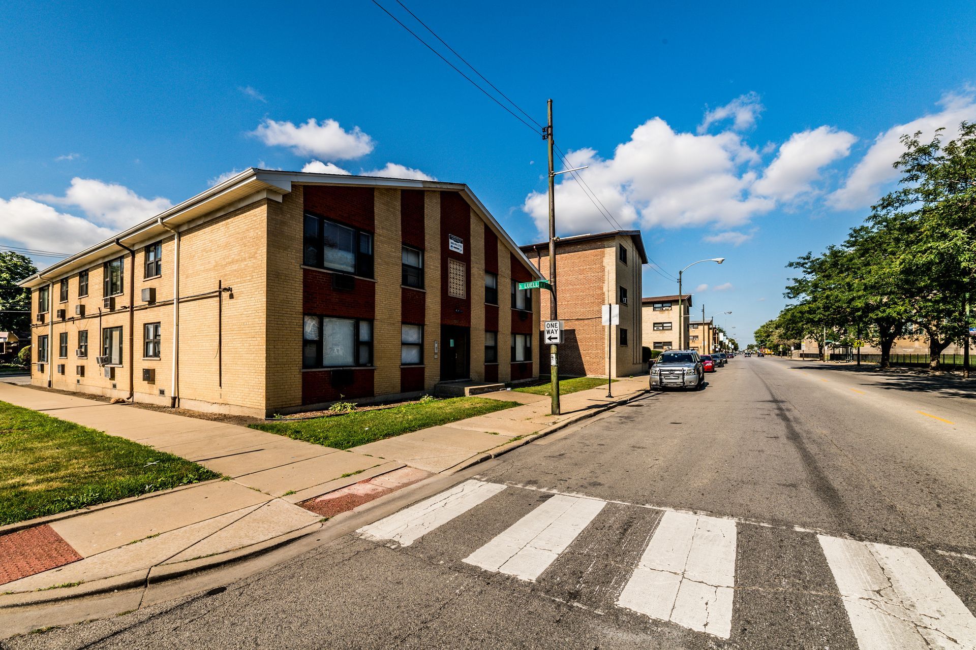 Apartment buildings on a city street with a crosswalk, blue sky, and a few parked cars.
