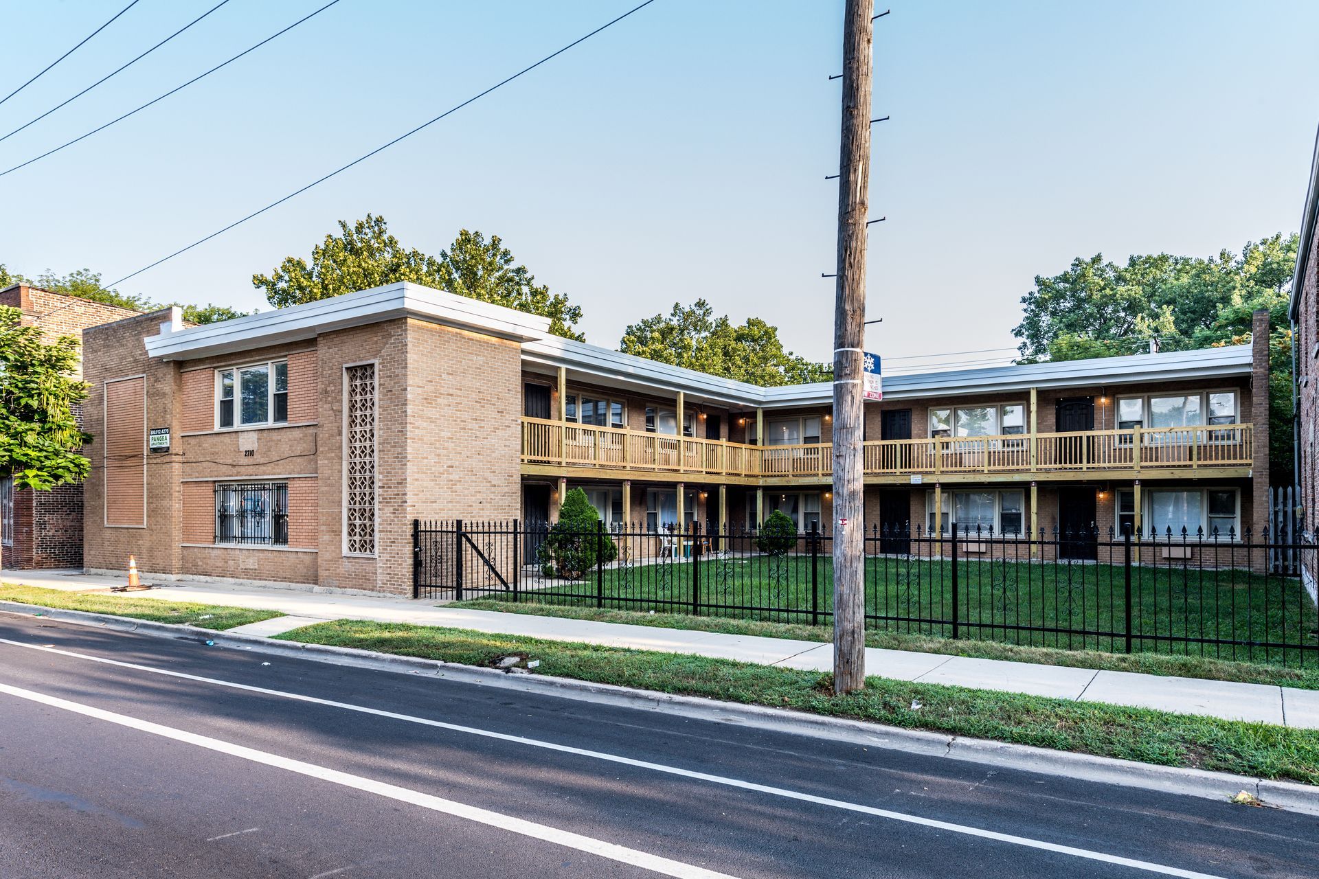 Two-story brick apartment building with balconies, black fence, and utility pole along a street.