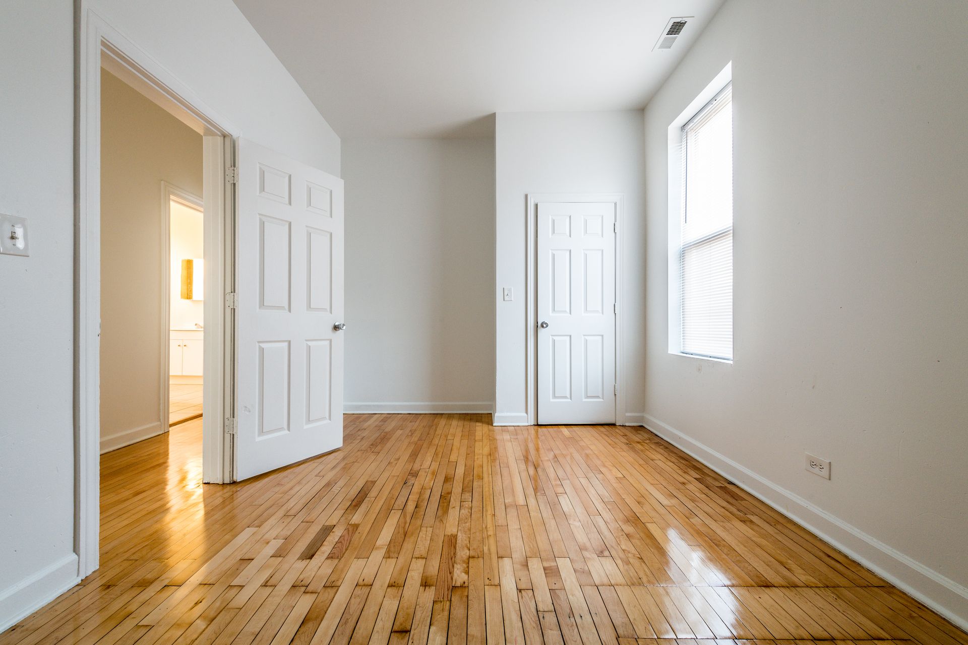 Empty room with hardwood floors, white walls, and three white doors. Window with blinds.