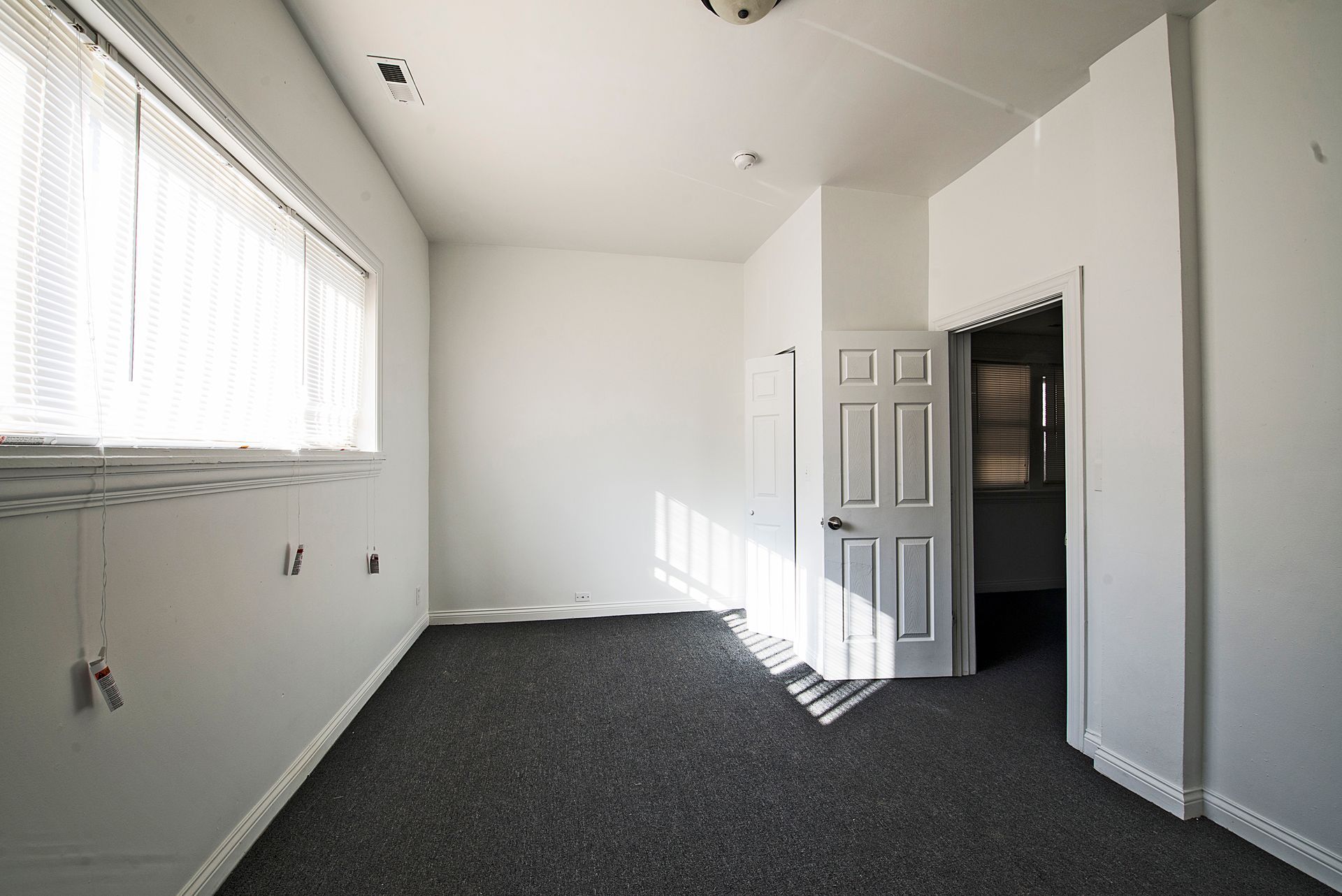 Empty room with white walls, dark carpet, and a window with blinds. Door to another room visible.
