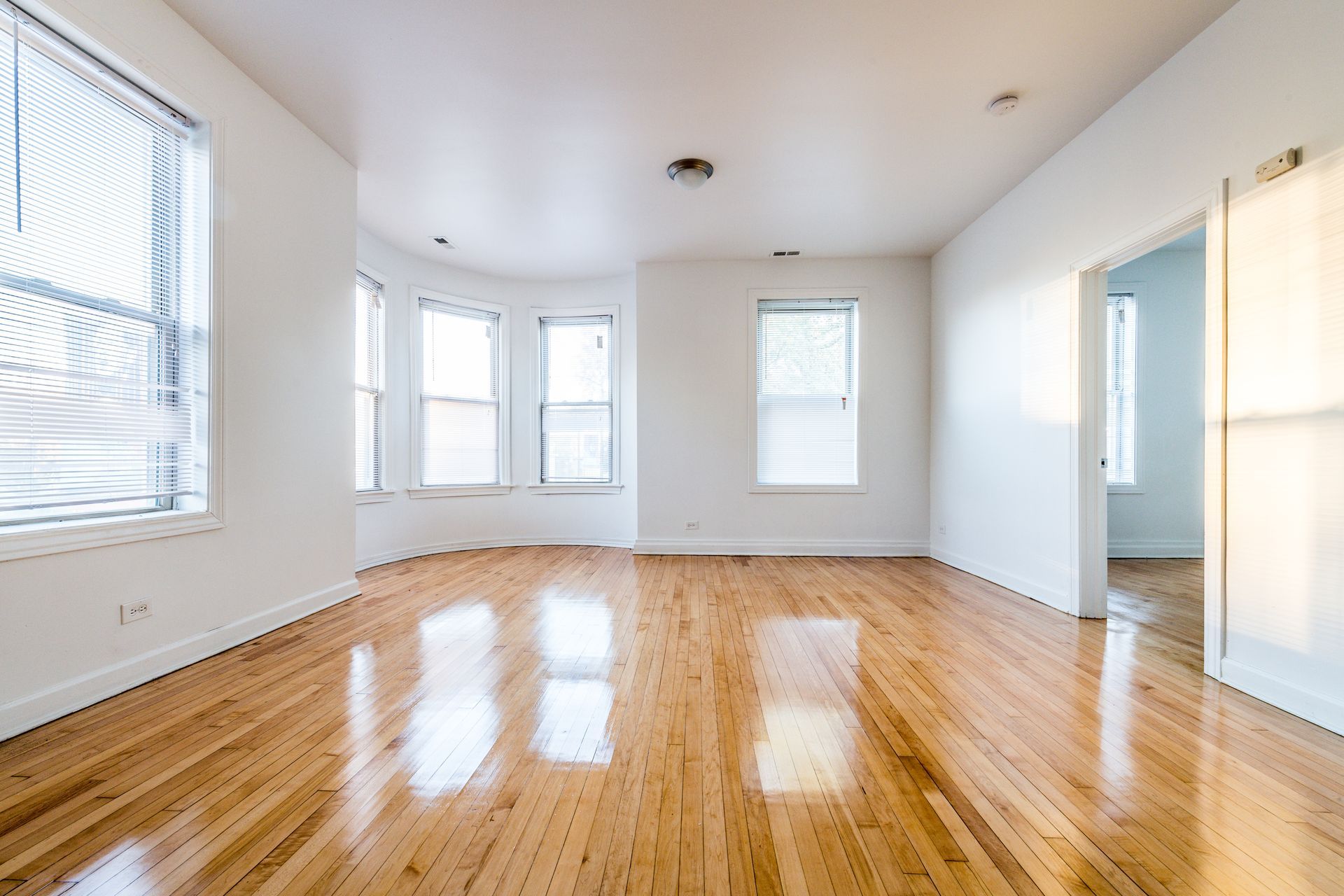 Empty room with hardwood floors, white walls, and bright sunlight through windows.
