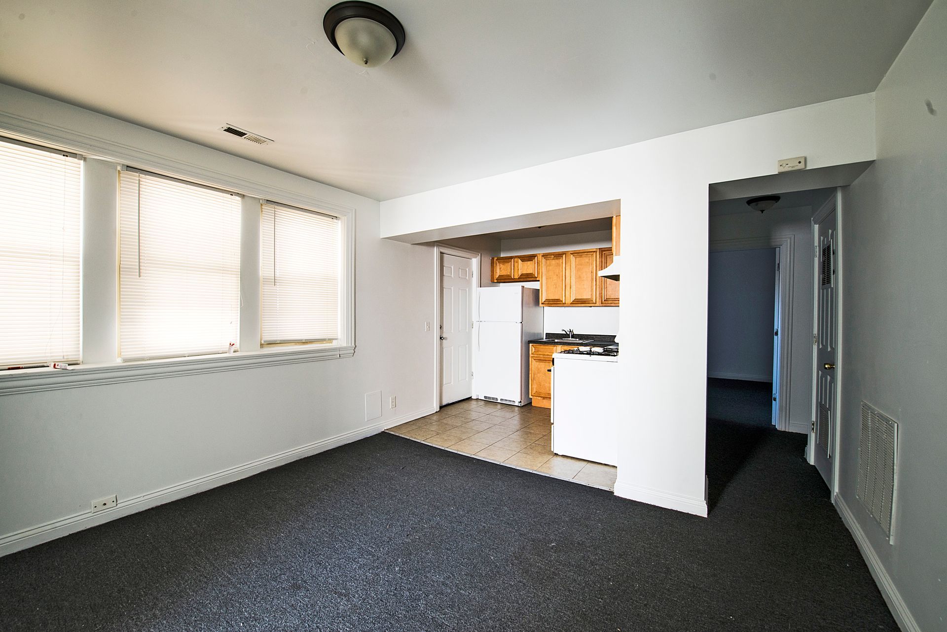 Empty apartment room with carpet, kitchen visible, and a hallway.