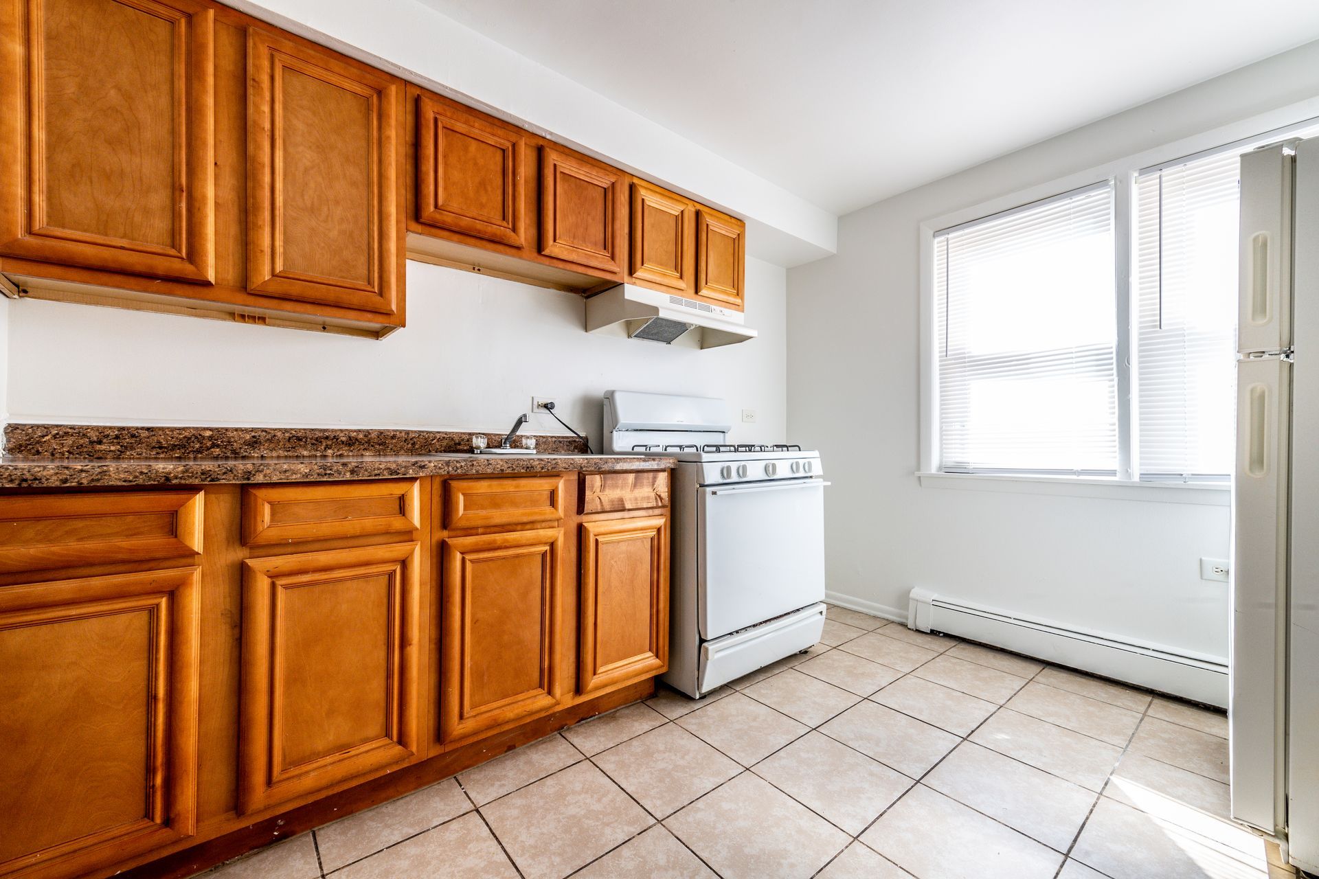 Kitchen with wooden cabinets, white appliances, and a window. Light-colored tile floor.