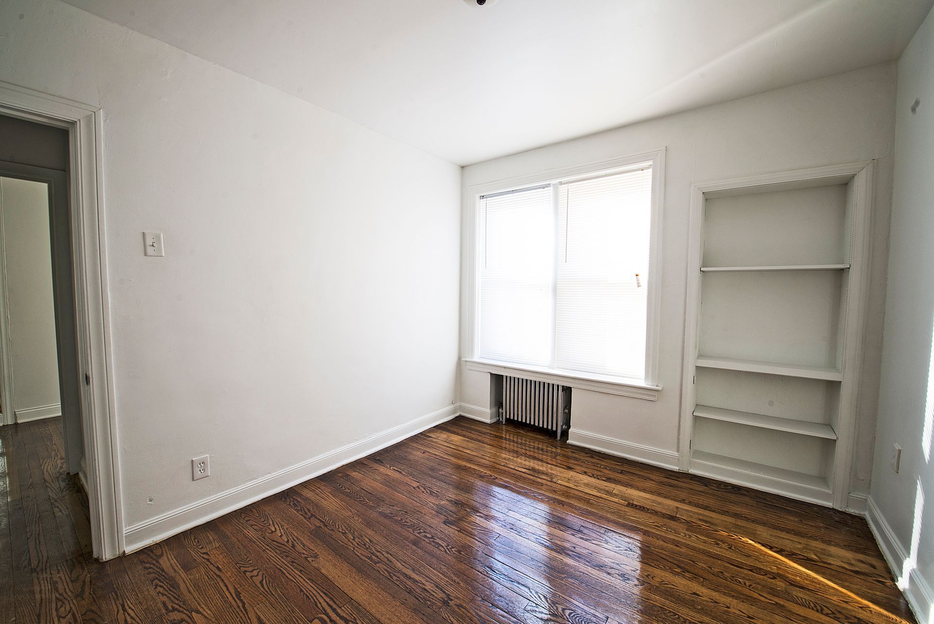 Empty room with hardwood floors, white walls, built-in shelves, and a window.