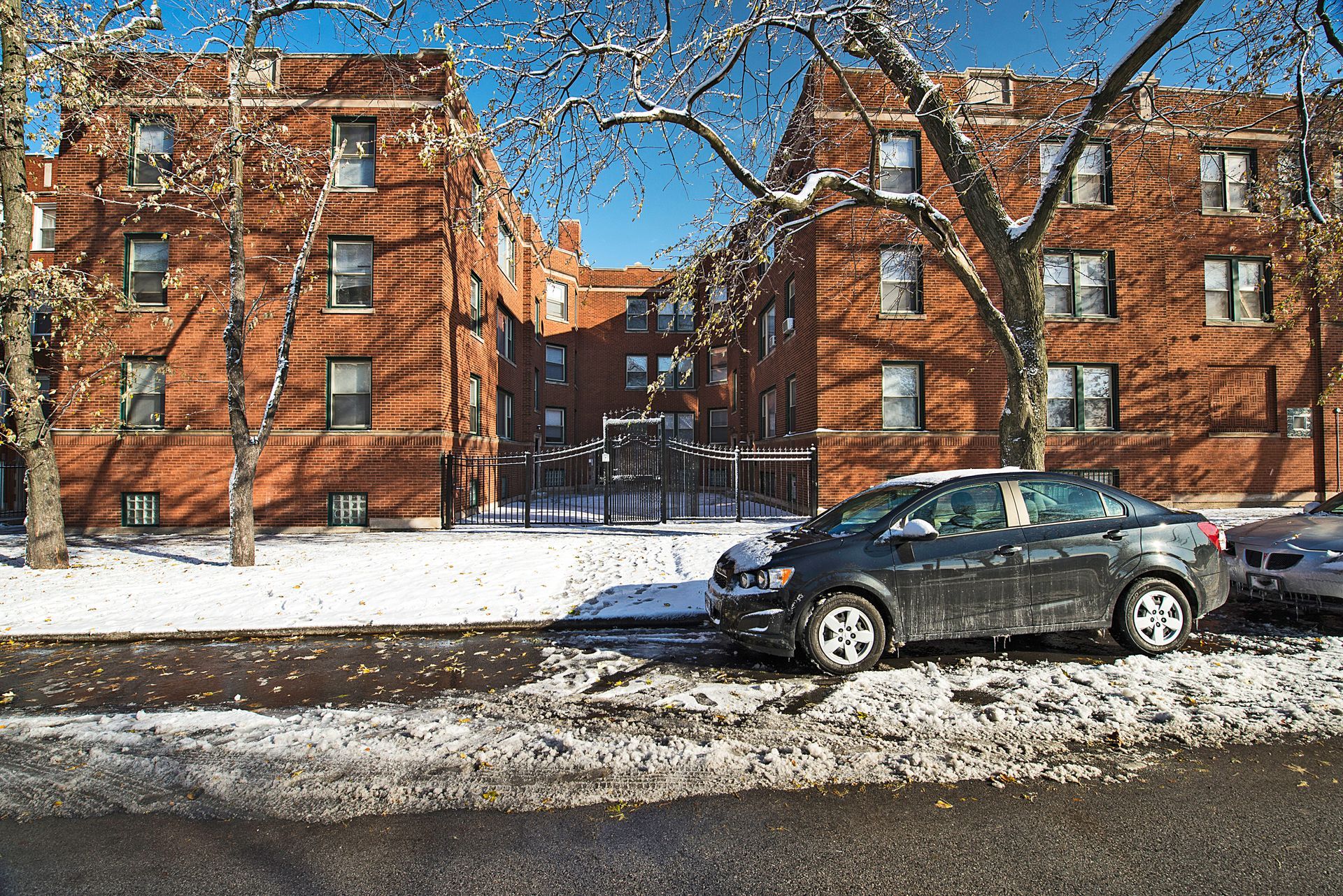 Snowy street scene with brick apartment buildings and parked cars.