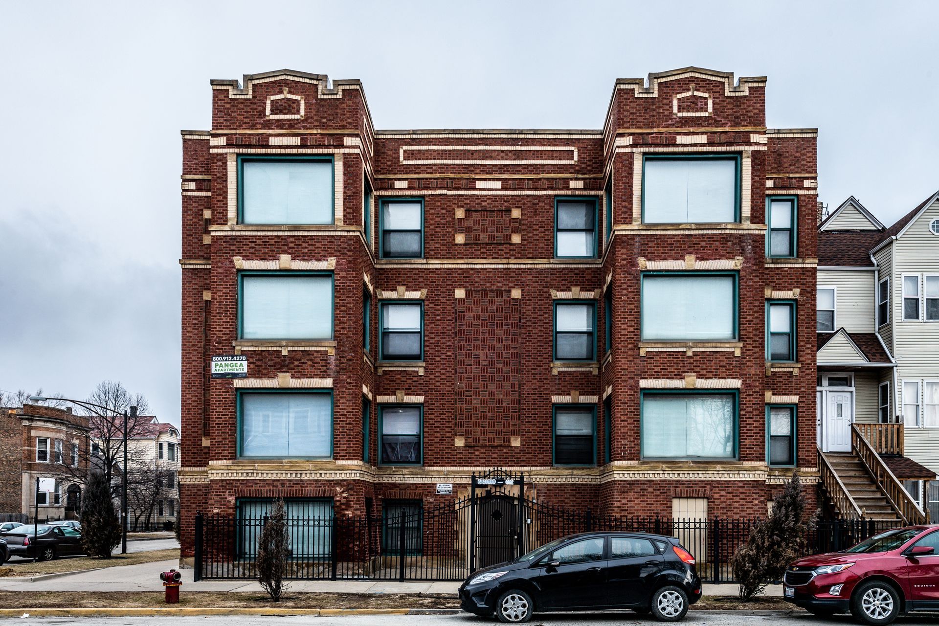 Brick apartment building with large windows and decorative trim on a cloudy day. Cars parked on the street.