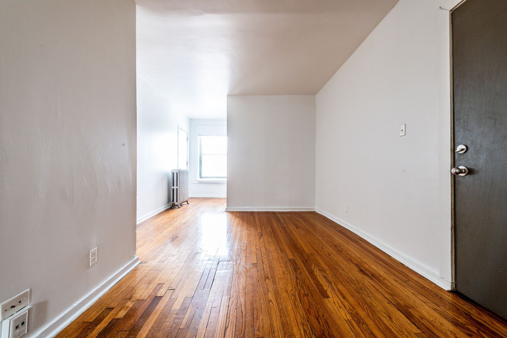 Empty room with hardwood floors, white walls, and a door on the right. Window at the end of the room.