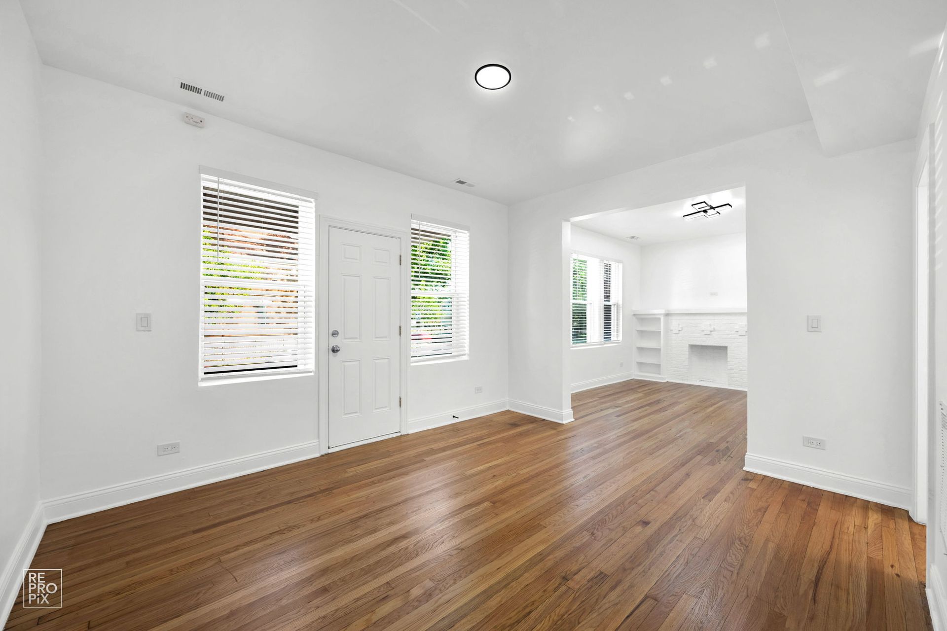 Empty room with hardwood floors, white walls, and a doorway leading to another room with a fireplace.