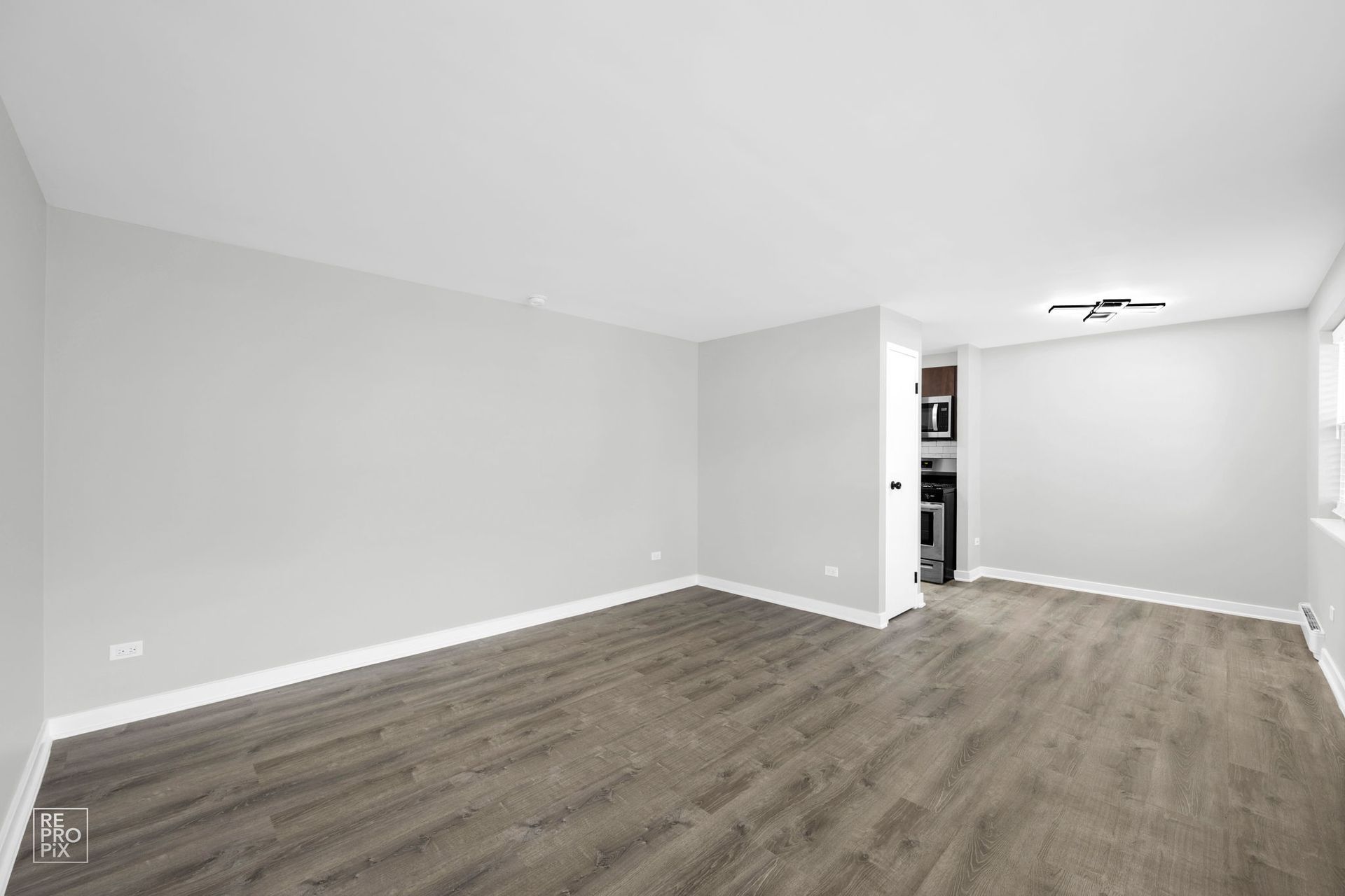 Empty living room with gray walls and dark wood-look flooring. Doorway to kitchen visible.