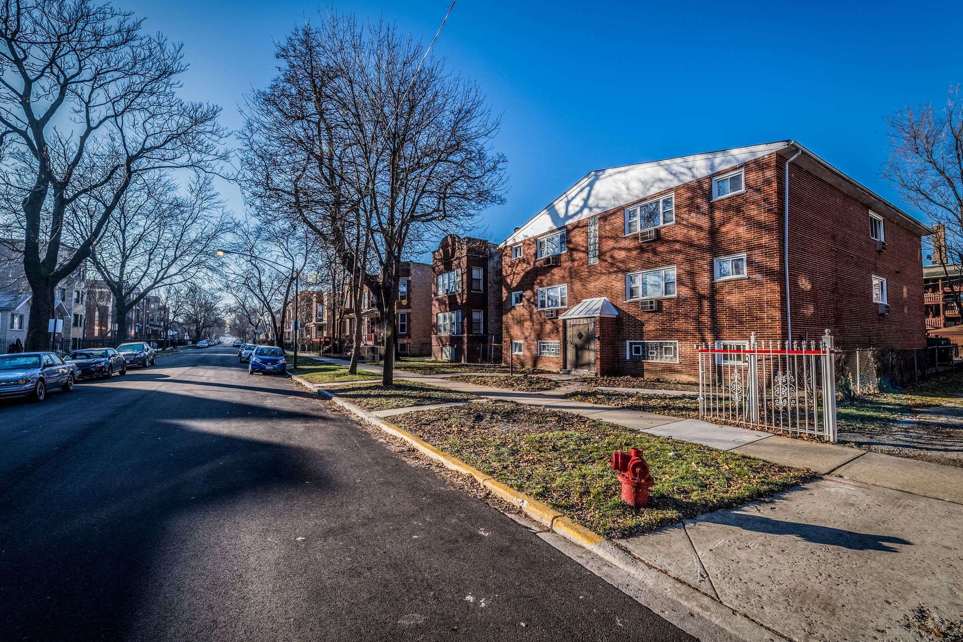 Street view of brick apartment buildings on a sunny day; cars parked along the curb.