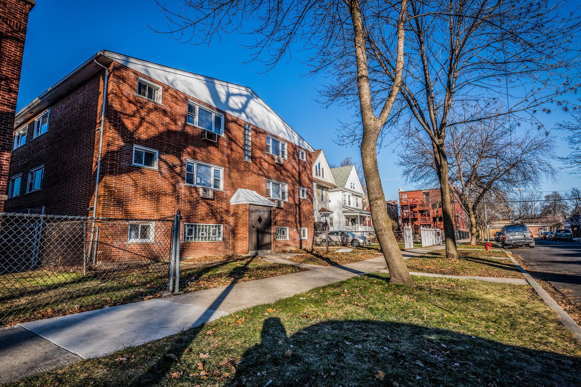 Red brick apartment building with sidewalk and street, sunny day. Bare trees.