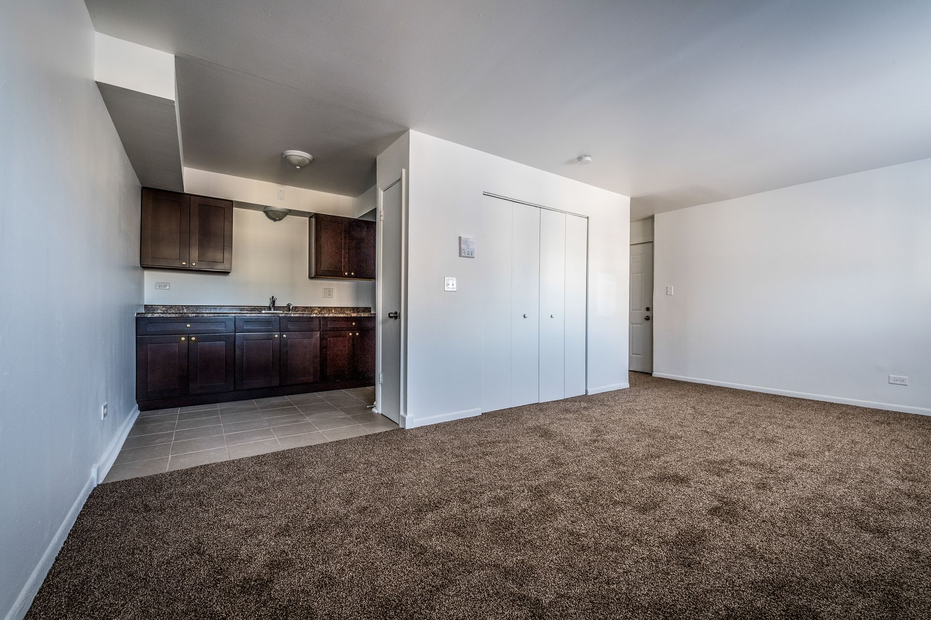 Empty apartment interior with brown carpet, dark cabinets, white walls, and closet doors.