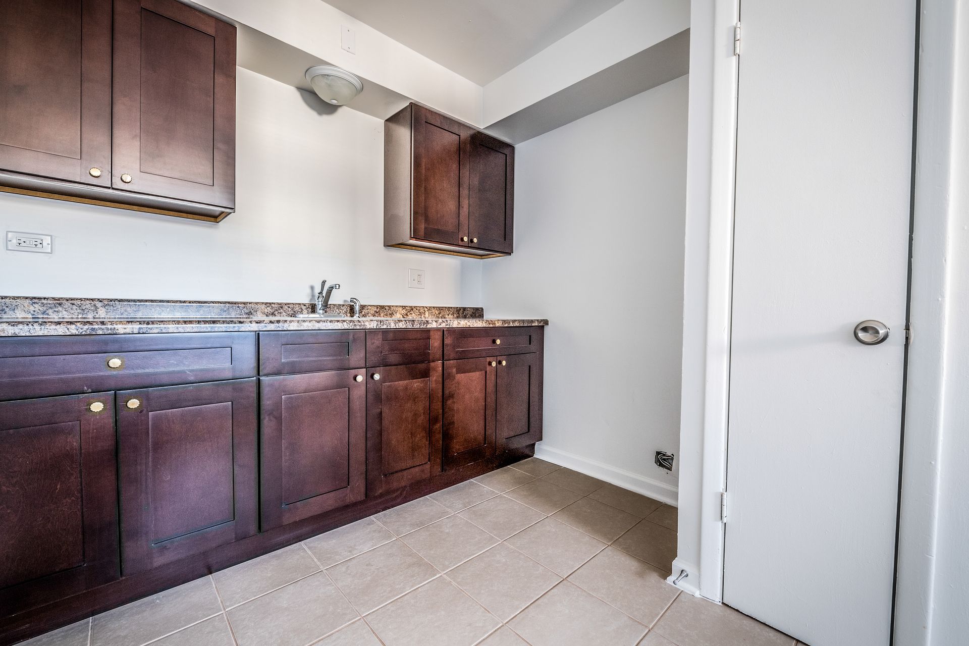 Kitchen with dark wood cabinets, granite countertop, sink, and tiled floor. Door on the right.