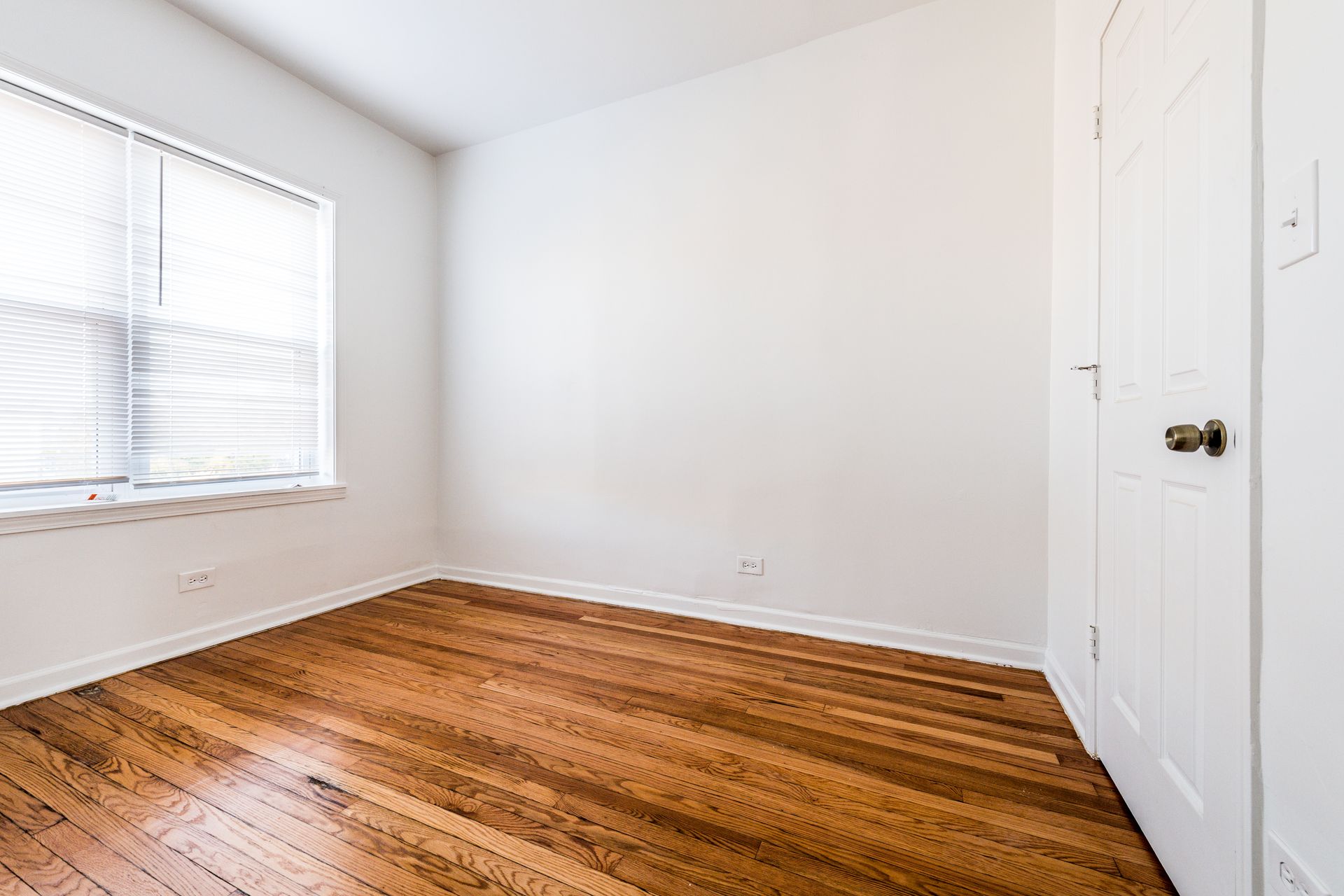 Empty room with hardwood floors, white walls, window with blinds, and a closed white door.