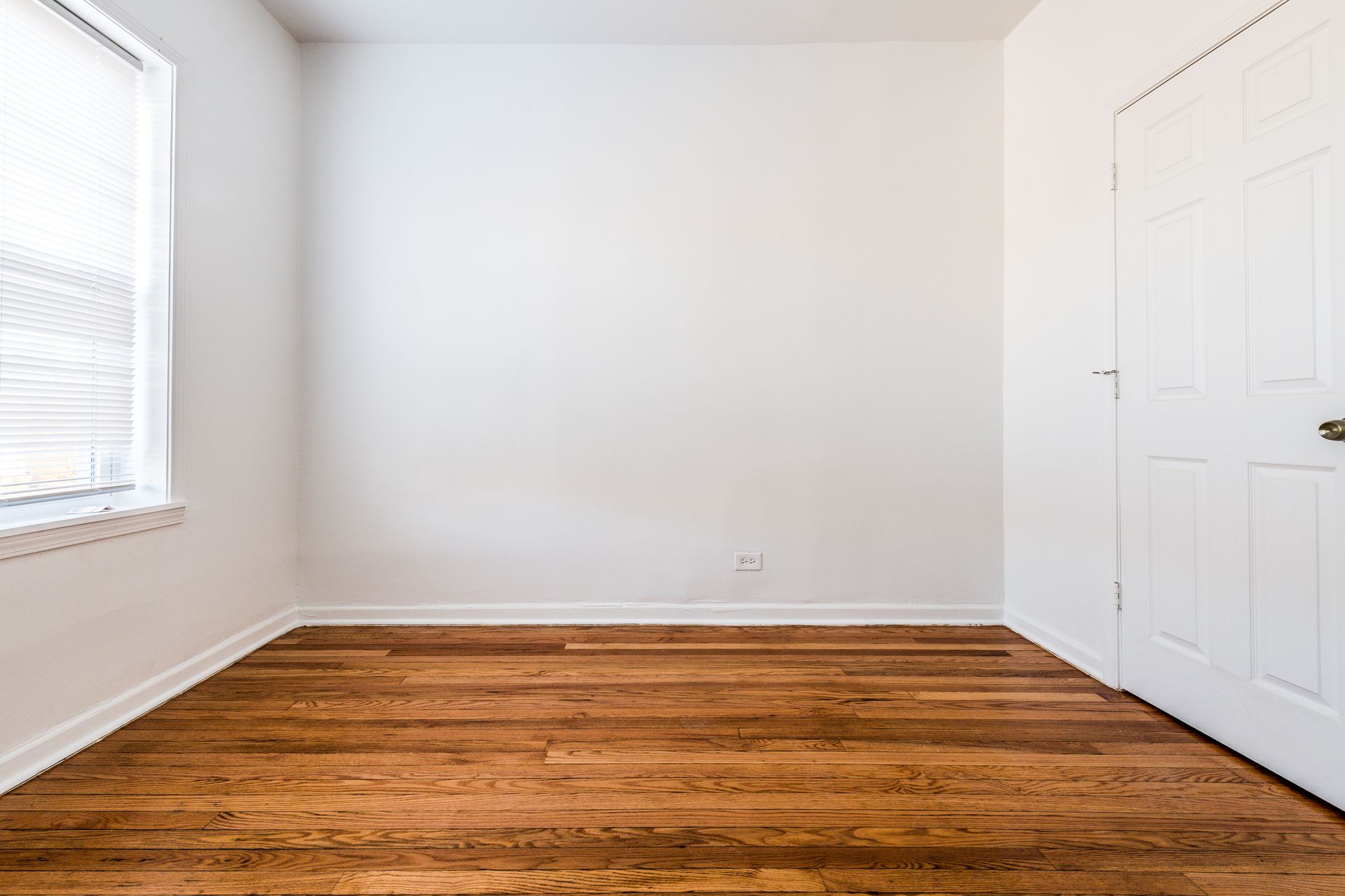 Empty room with hardwood floors, white walls, a window with blinds, and a closed door.