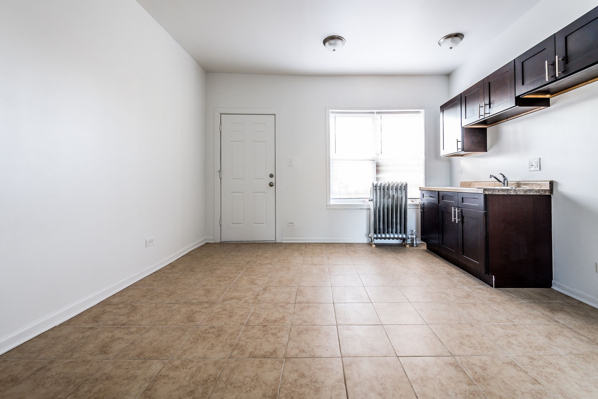 Empty room with kitchenette: white walls, brown cabinets, tile floor, window, and door.